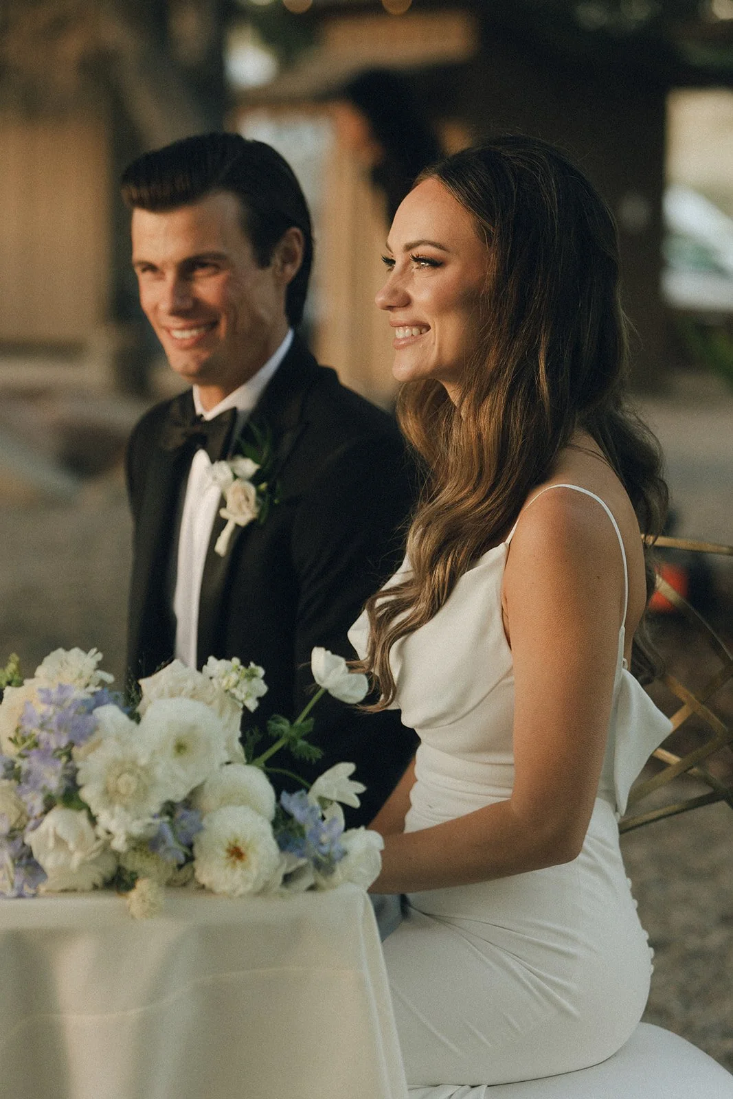 A bride and groom sitting at a table during their wedding celebration, smiling and looking to the side. The bride has long, wavy hair and is wearing a white dress with thin straps. The groom has dark hair and is dressed in a black tuxedo with a white