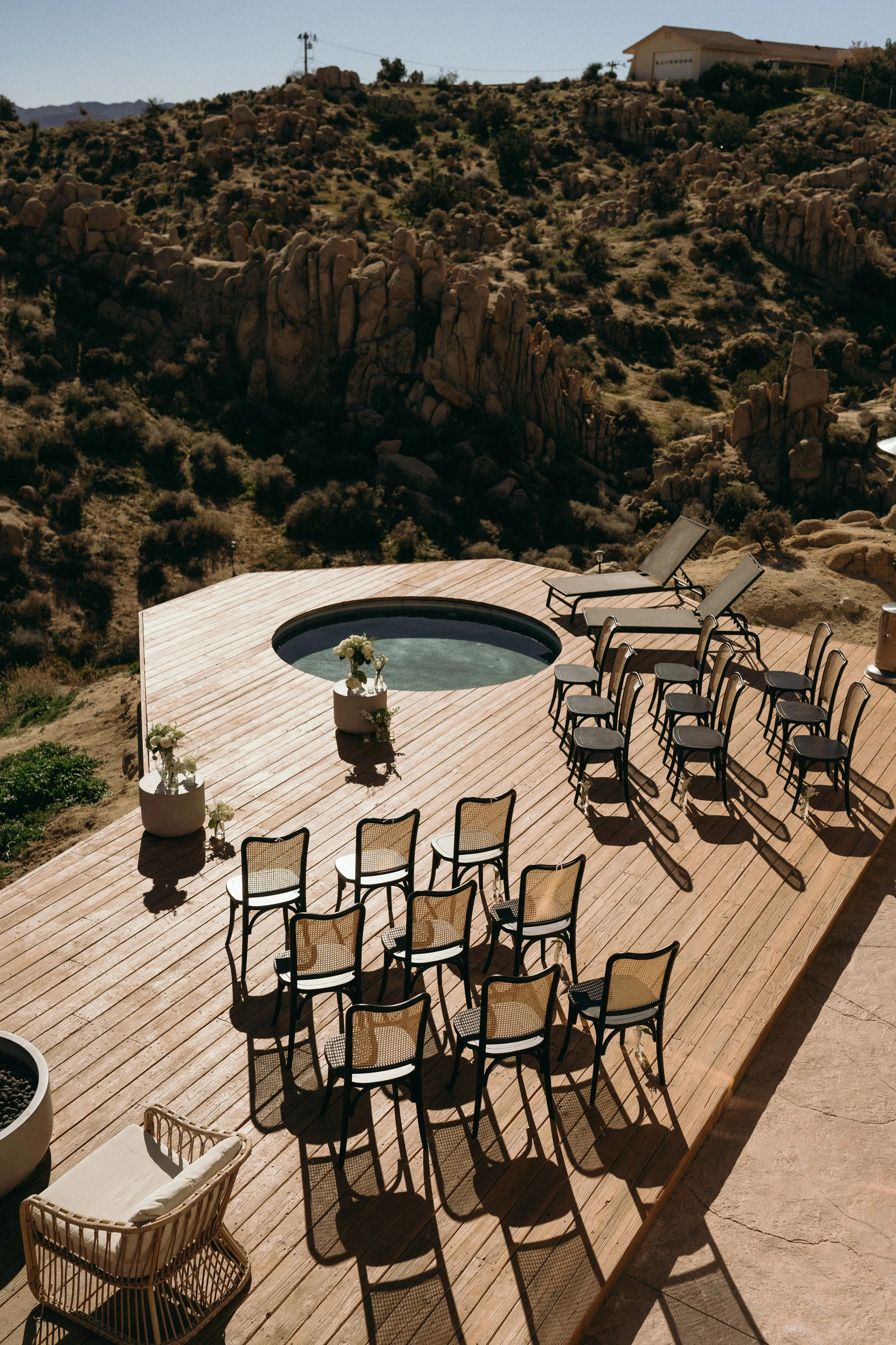 An outdoor wooden deck with chairs arranged for a gathering, two sun loungers, and a small hot tub, with a rocky, mountainous landscape in the background.
