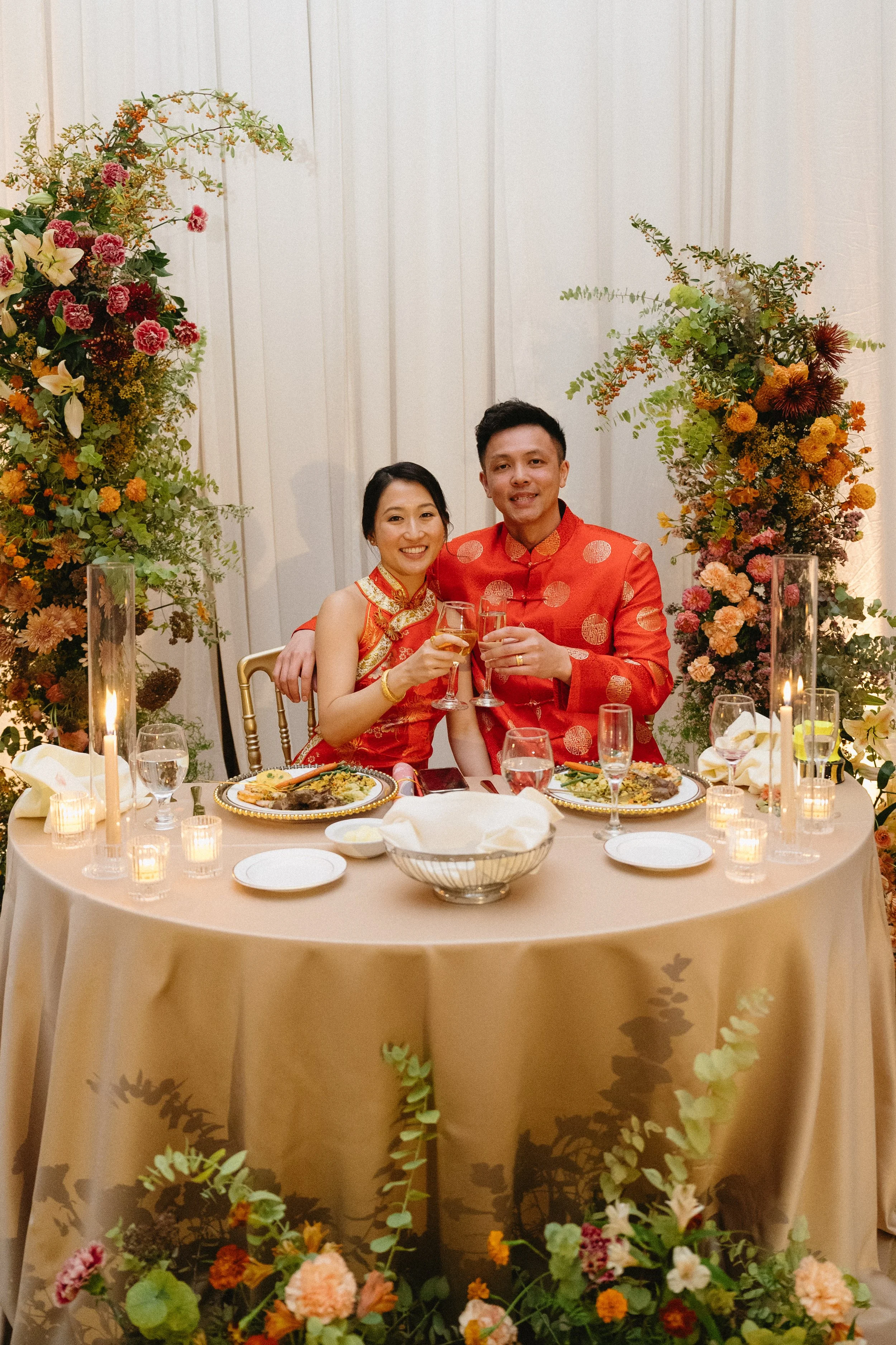 A couple dressed in traditional Chinese red wedding attire celebrating their wedding at a decorated banquet table with flowers and candles.