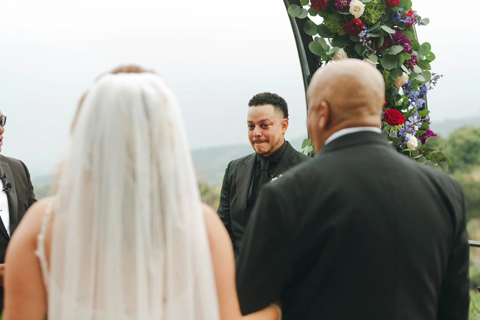 A groom with short curly hair and a goatee, dressed in a black suit, stands during a wedding ceremony outdoors, facing a bride with long blonde hair and a veil, and an officiant. The scene is decorated with colorful flowers on an arch, with a scenic