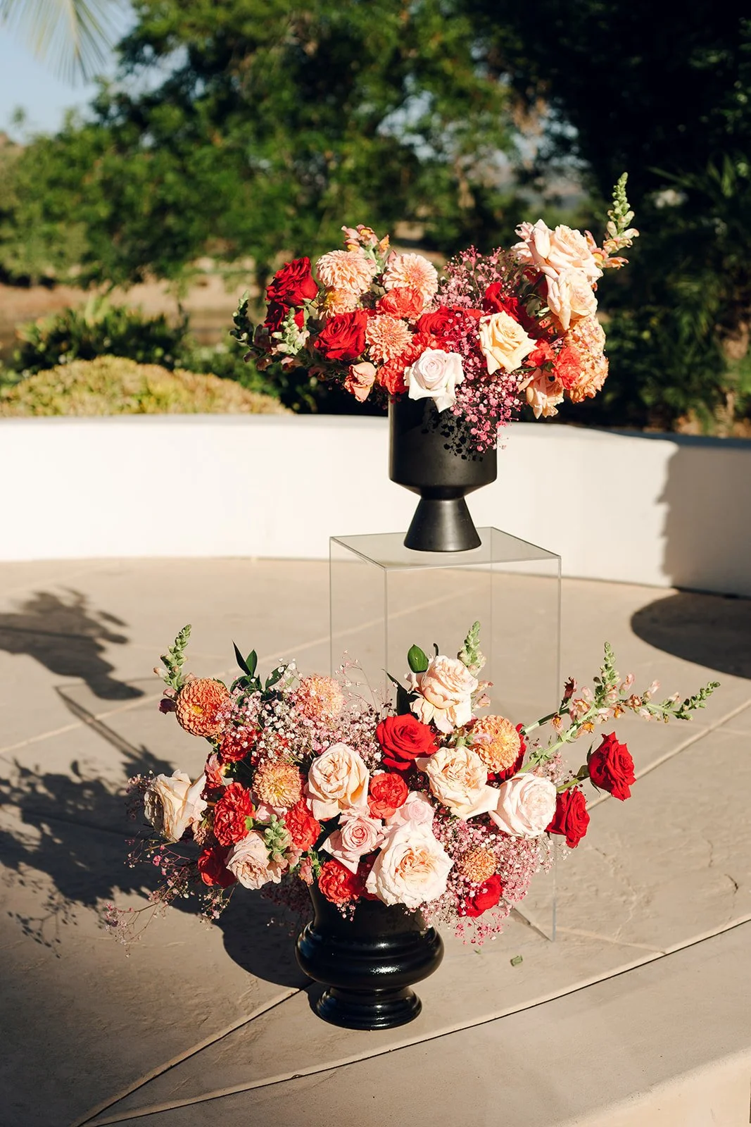 Two floral arrangements with pink, red, and white roses, placed in black vases, with one on a clear acrylic stand outdoors in sunlight.