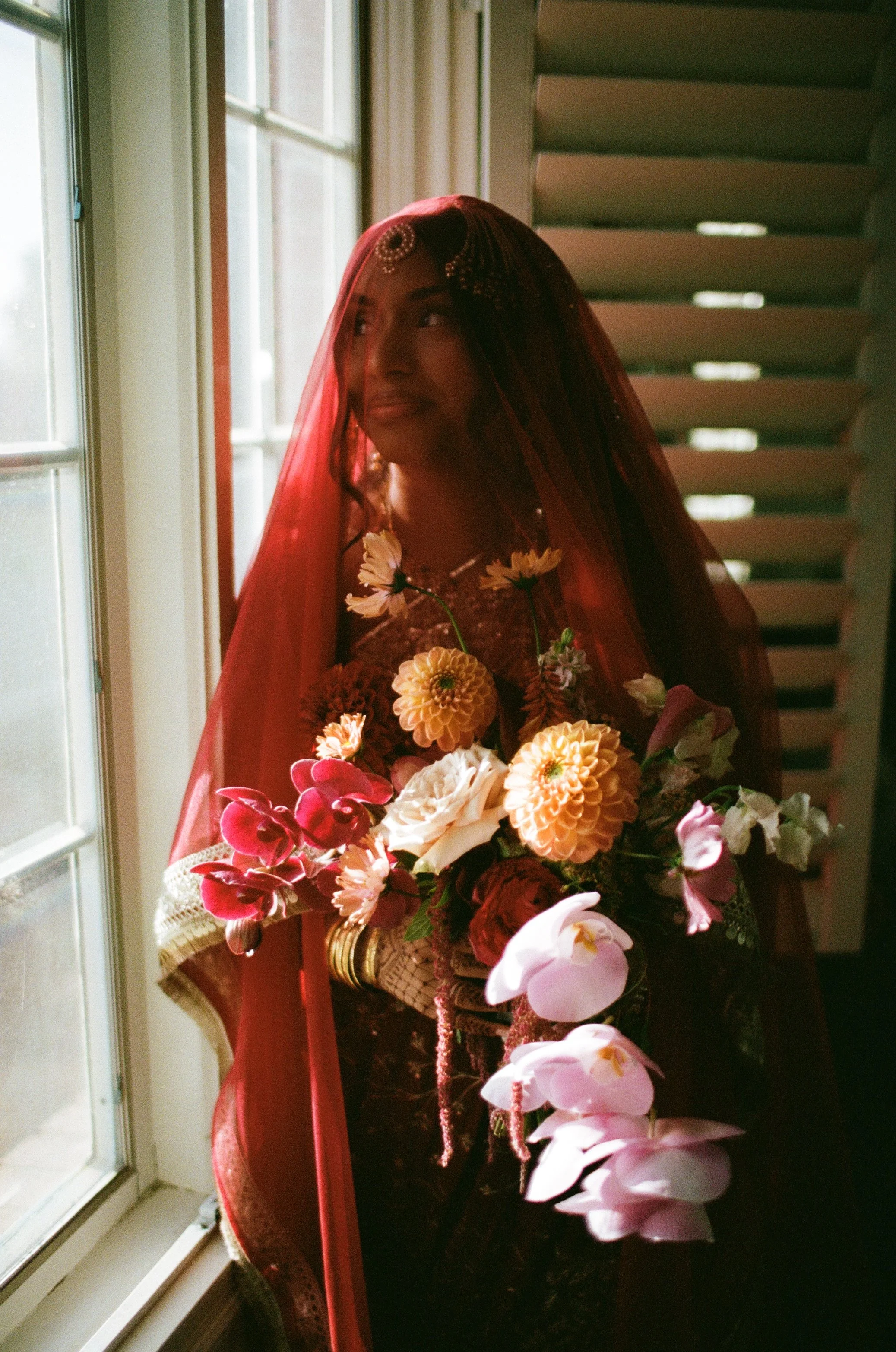 A woman dressed as a bride wearing a red veil and jewelry, holding a bouquet of assorted flowers, standing near a window inside a house.
