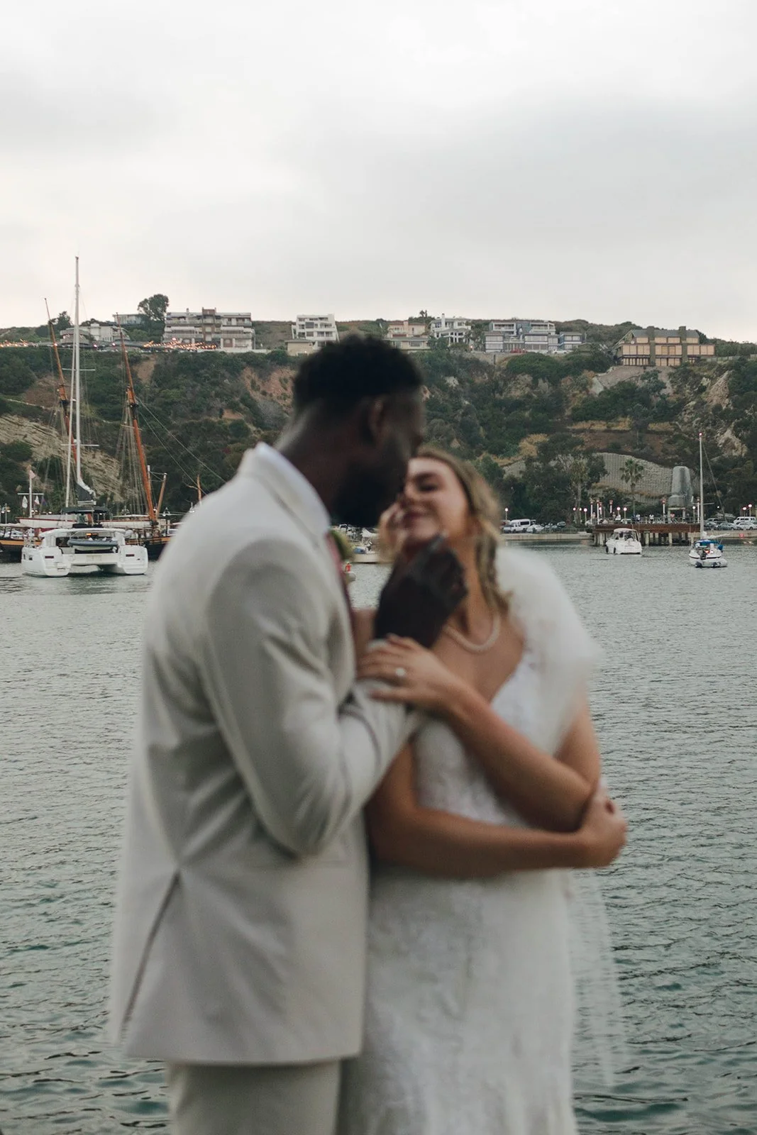 A couple in wedding attire sharing an intimate moment by the water with boats and a hillside with houses in the background.