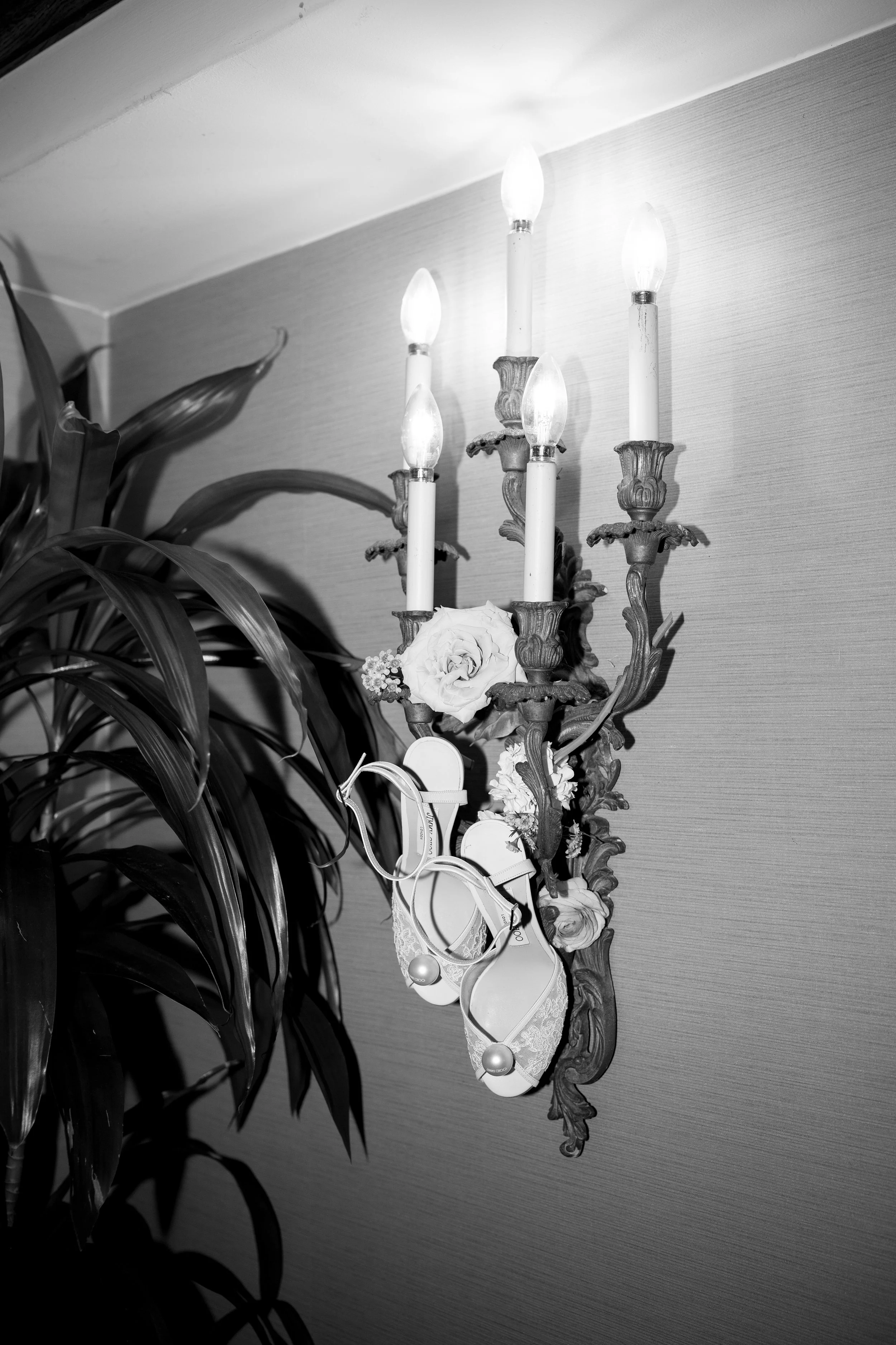 A creative shot of the bride's Jimmy Choo wedding heels hanging from the candelabra style lamp in the hotel hallway