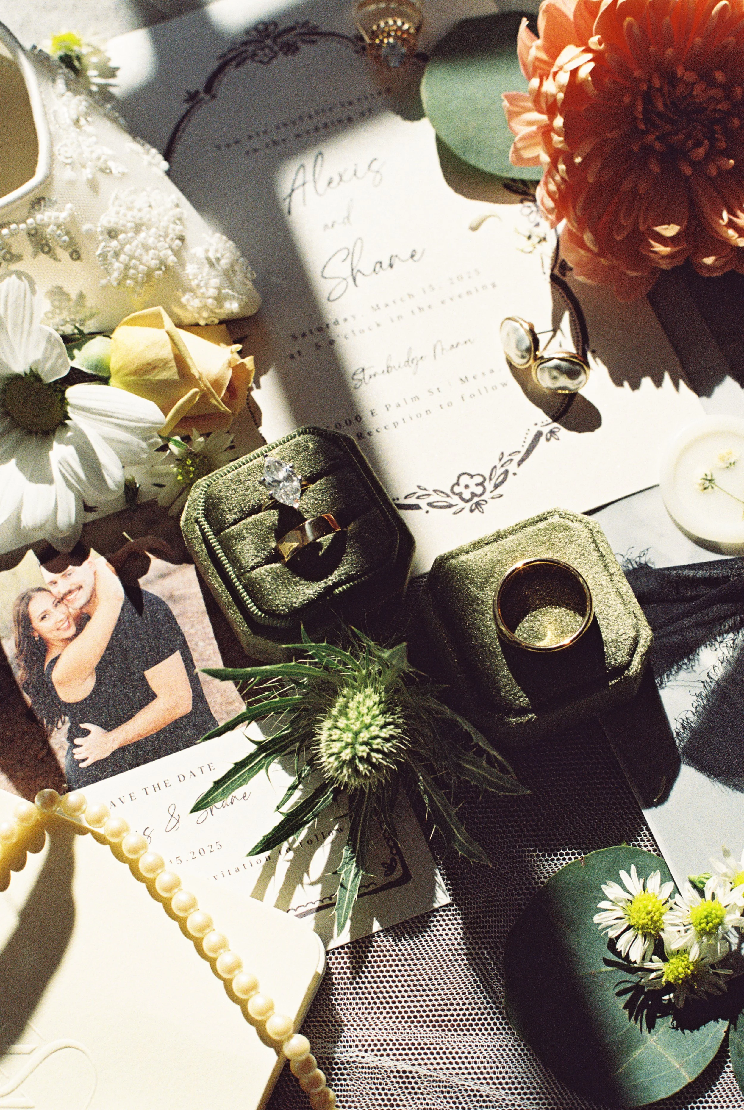 A wedding invitation, engagement ring, wedding bands, flowers, pearl necklace, and candles arranged on a table.