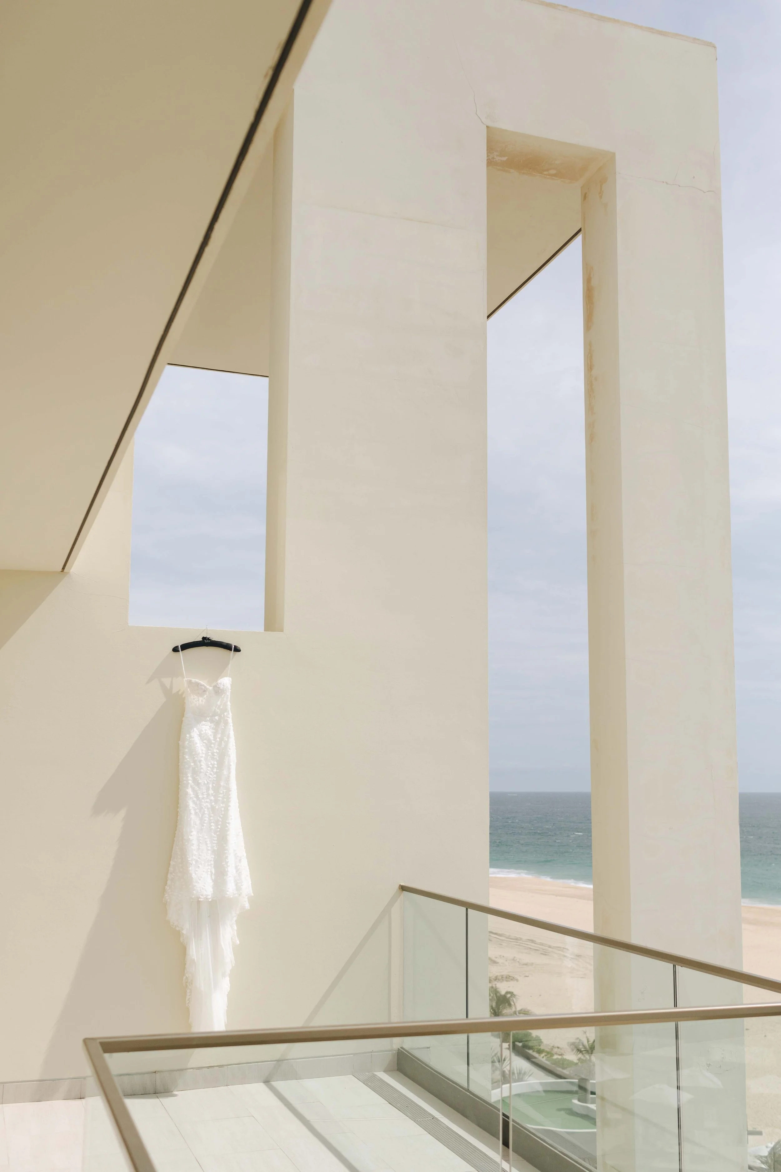 A white wedding dress hanging on a black hanger on a pale yellow wall inside a modern building overlooking the beach and ocean.