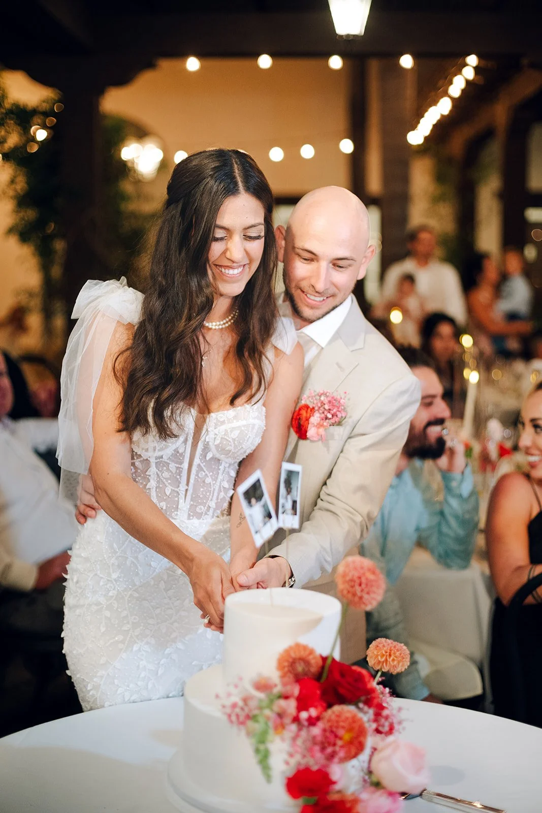 A bride and groom cutting their wedding cake, smiling, in a decorated reception hall with guests in the background.