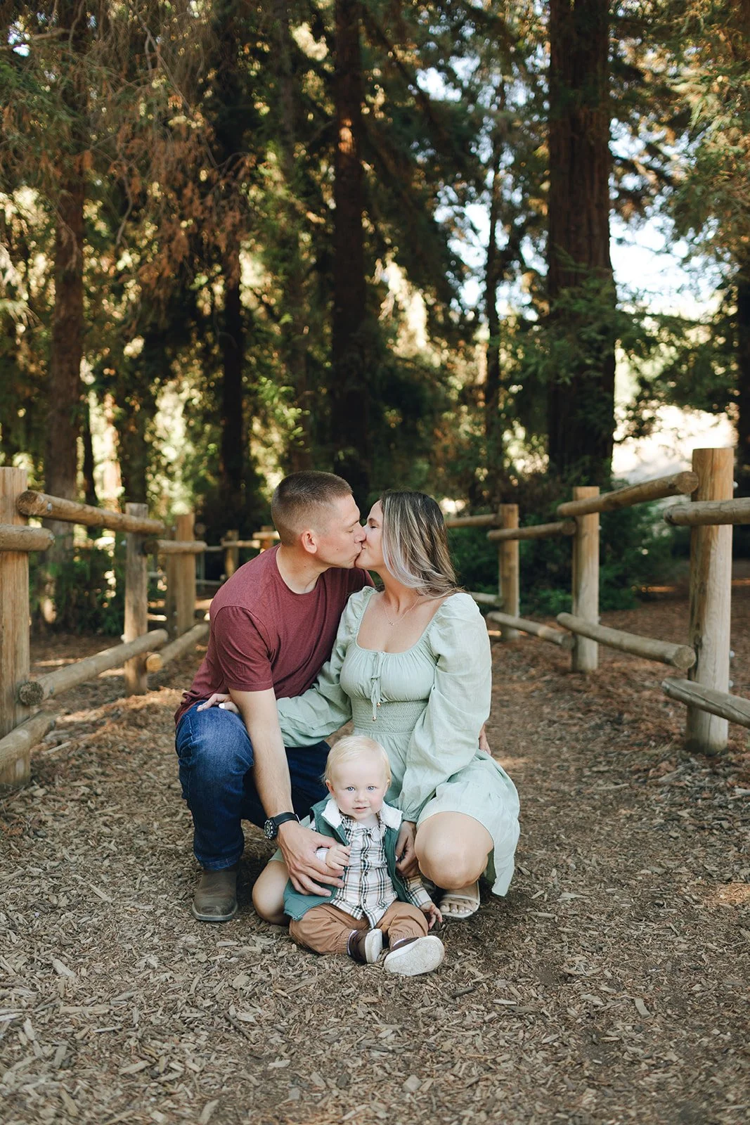 A family of three sitting on a wooded trail, kissing each other. The father and mother are sharing a kiss while the young child is sitting in front of them, smiling at the camera. Tall trees line the background.