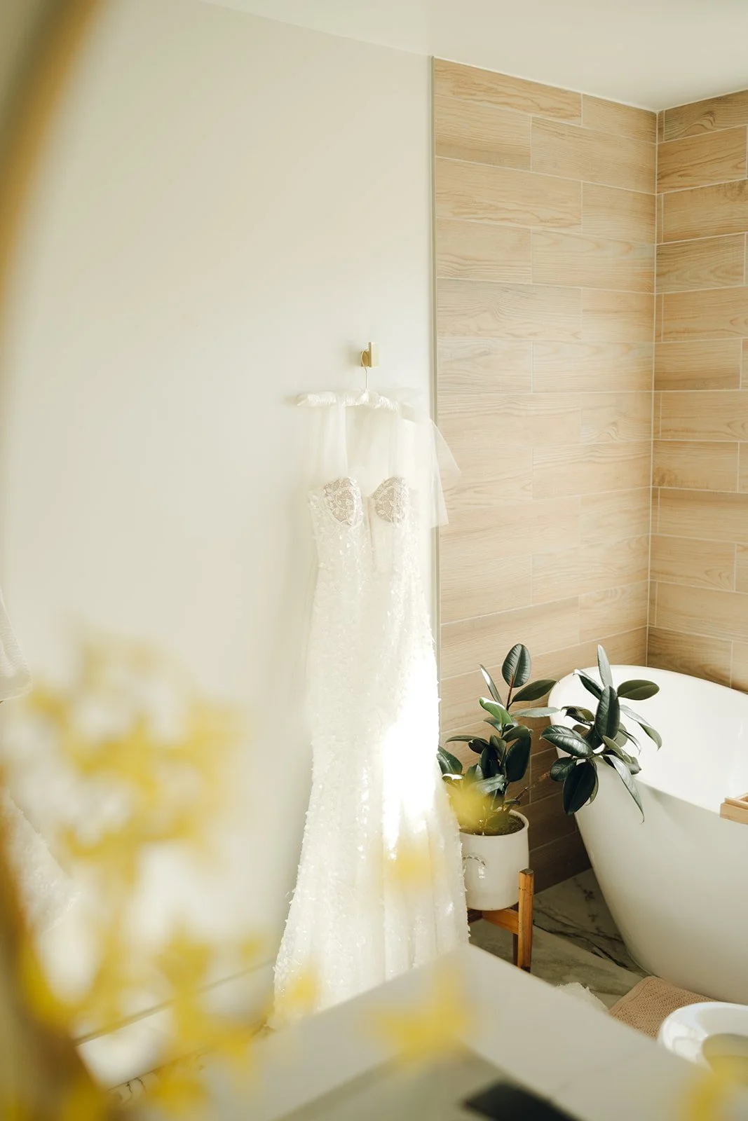 White wedding dress hanging on a wall hook in a bathroom with beige tiled walls and a modern bathtub, next to a potted plant.