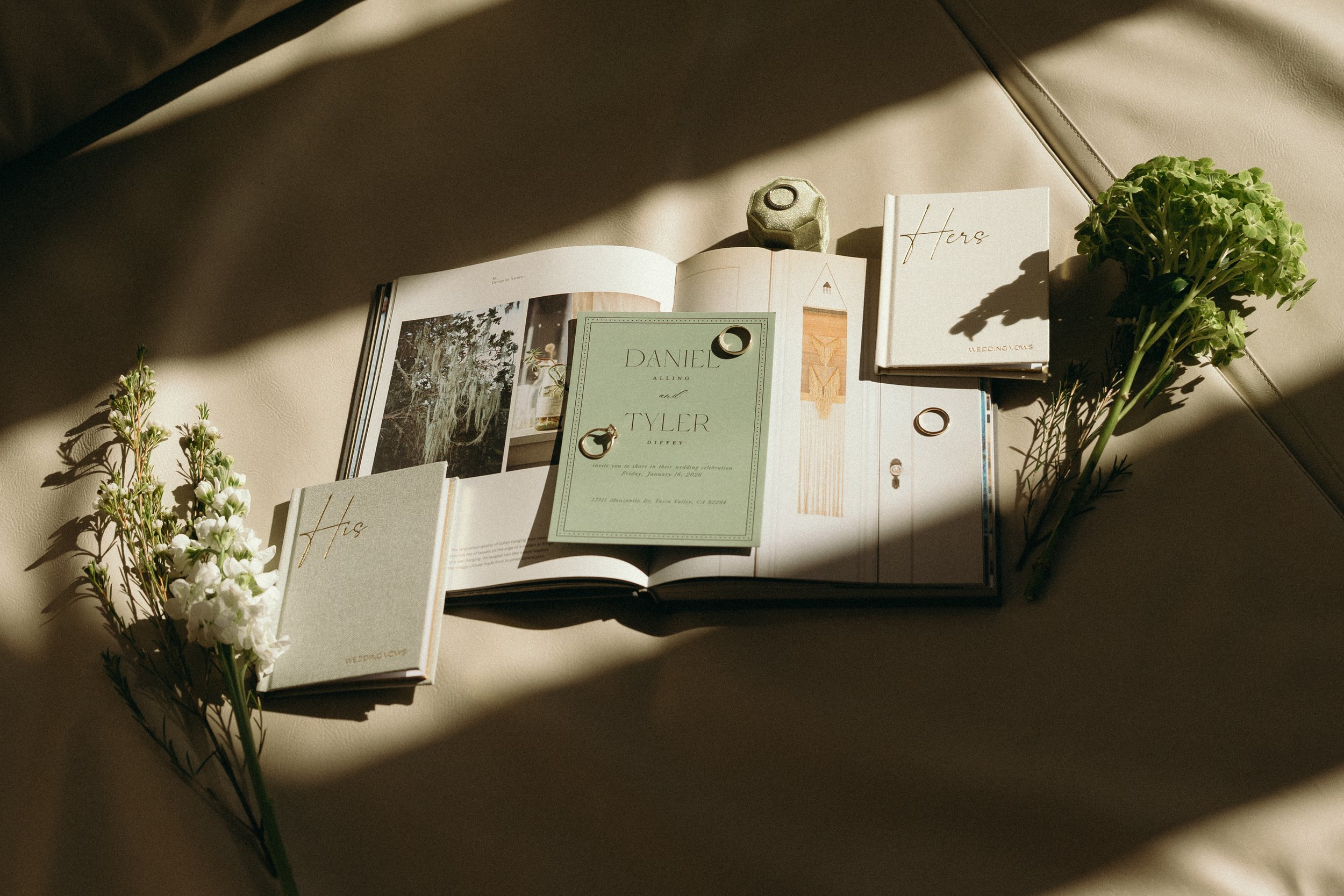 Open wedding photo album, wedding invitations, small closed albums labeled 'His' and 'Hers', white flowers, and a small gray decorative object on a cream leather surface, with sunlight casting shadows.