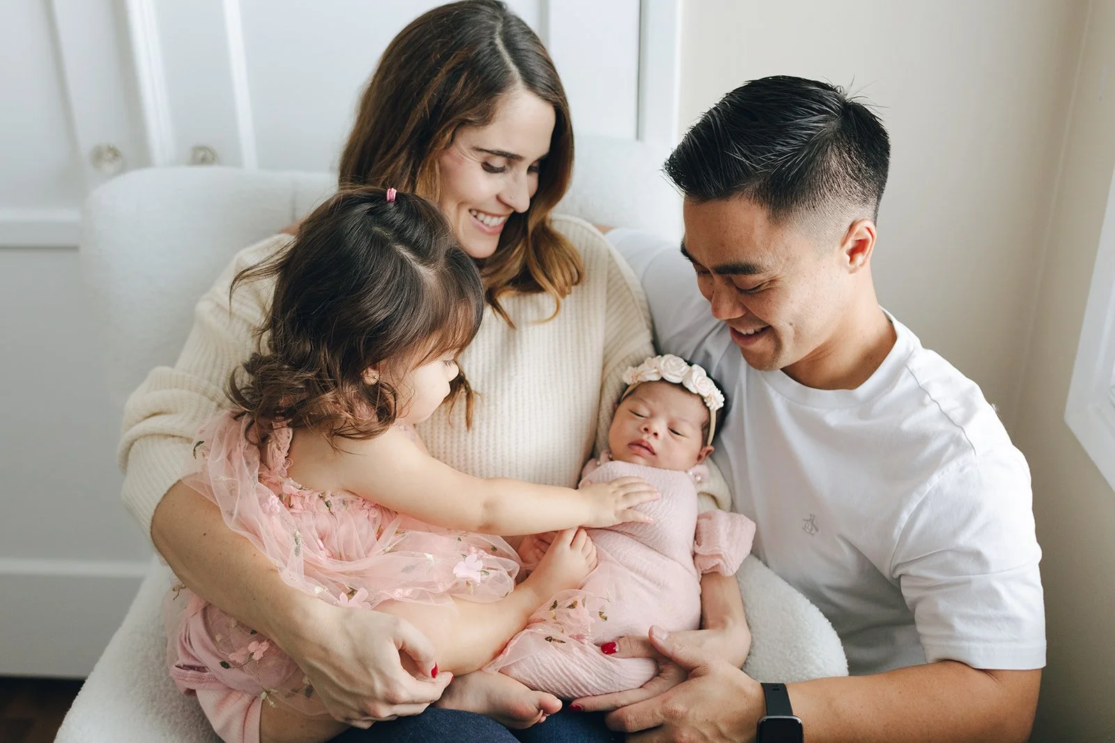 Family of four, including a newborn, sitting on a white couch in a bright room, smiling and interacting with each other.