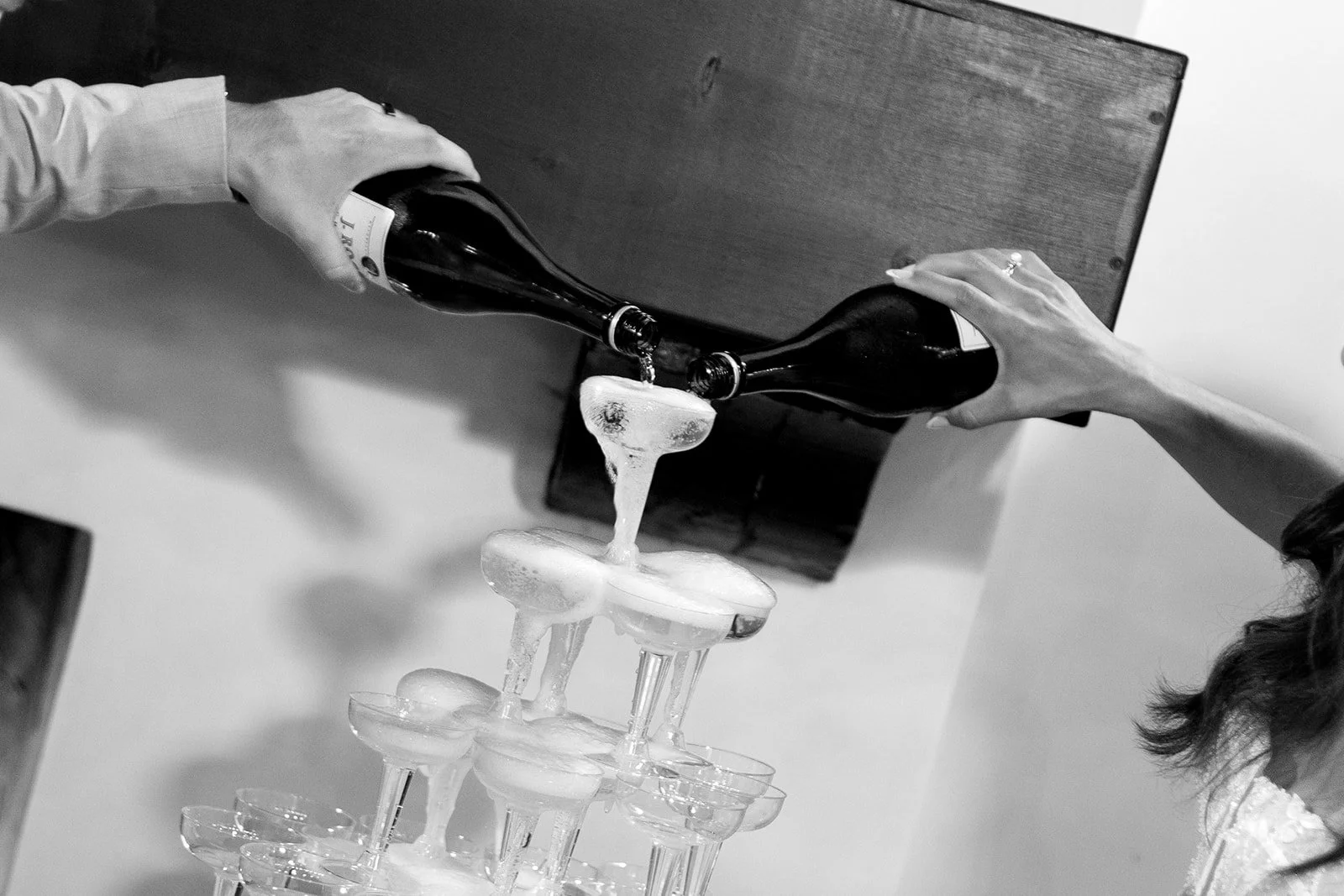 Two people are pouring champagne bottles into a pyramid of glasses on a table.