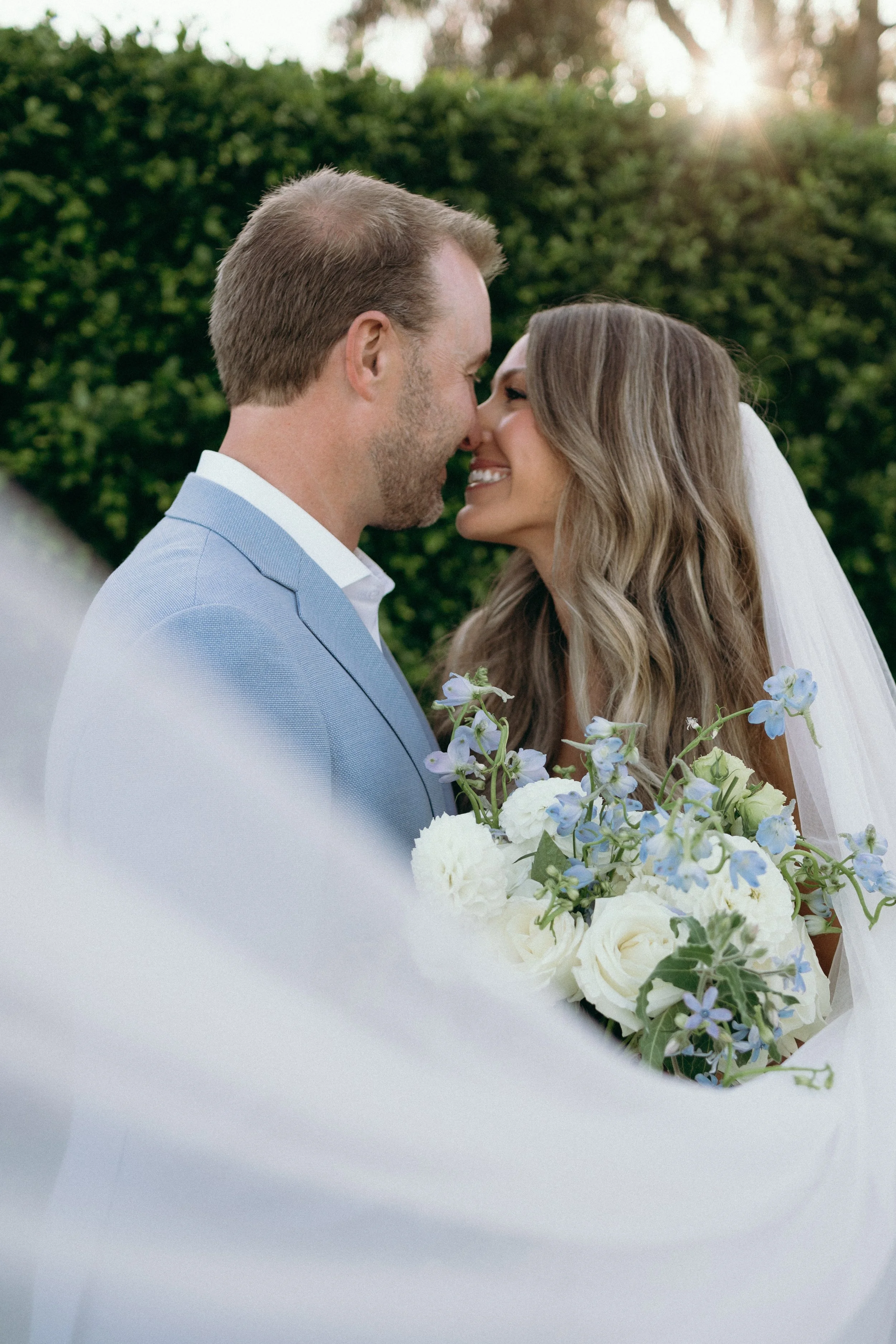 A happy bride and groom on their wedding day, touching foreheads and smiling outdoors with greenery and sunlight in the background, the bride holding a bouquet of white and blue flowers.