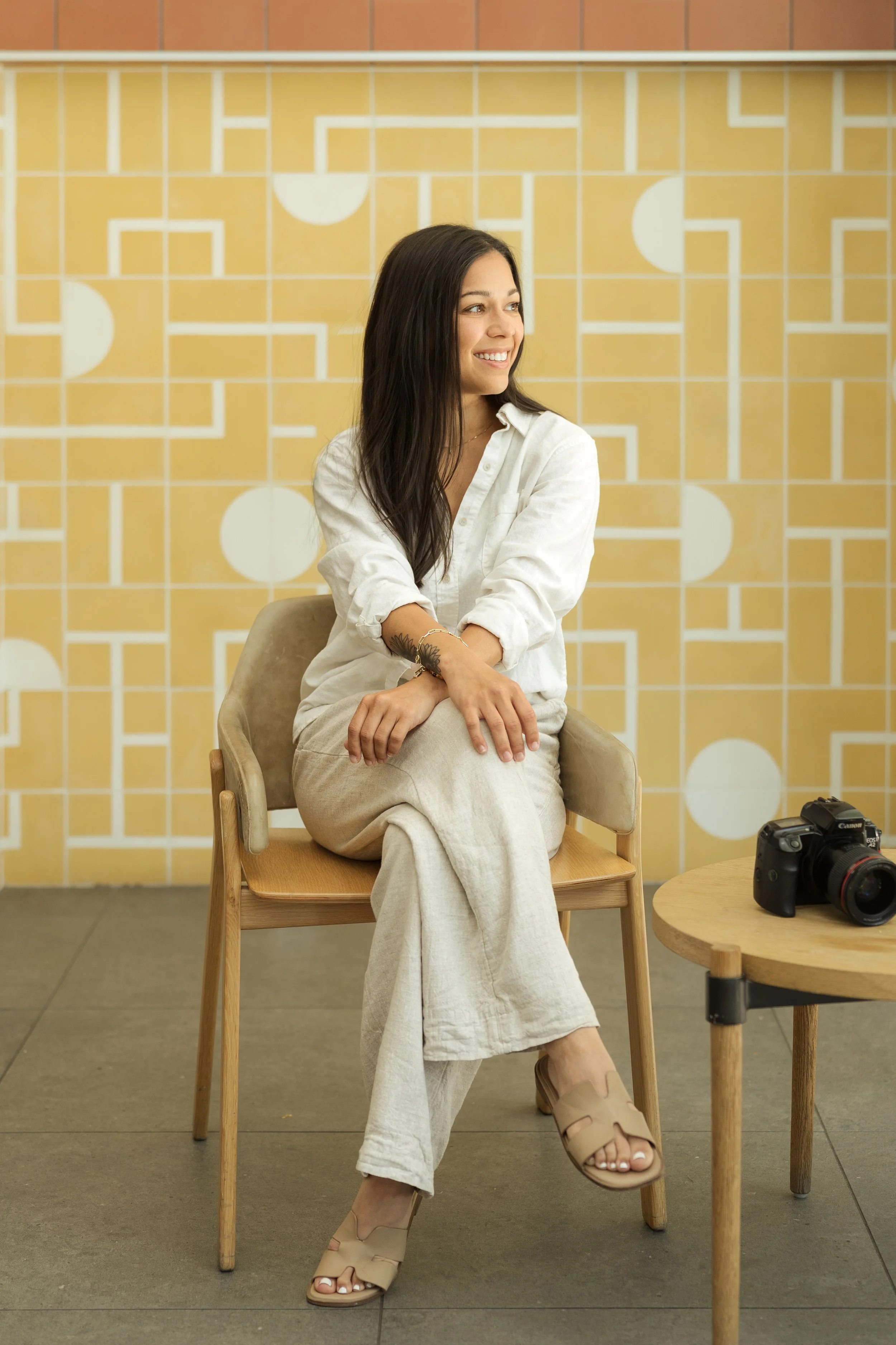 A woman with long dark hair, wearing a white shirt and beige pants, sitting on a wooden chair with her legs crossed and smiling. There is a camera on a round wooden table to her right. The background features a yellow and white tiled wall with geometric patterns.