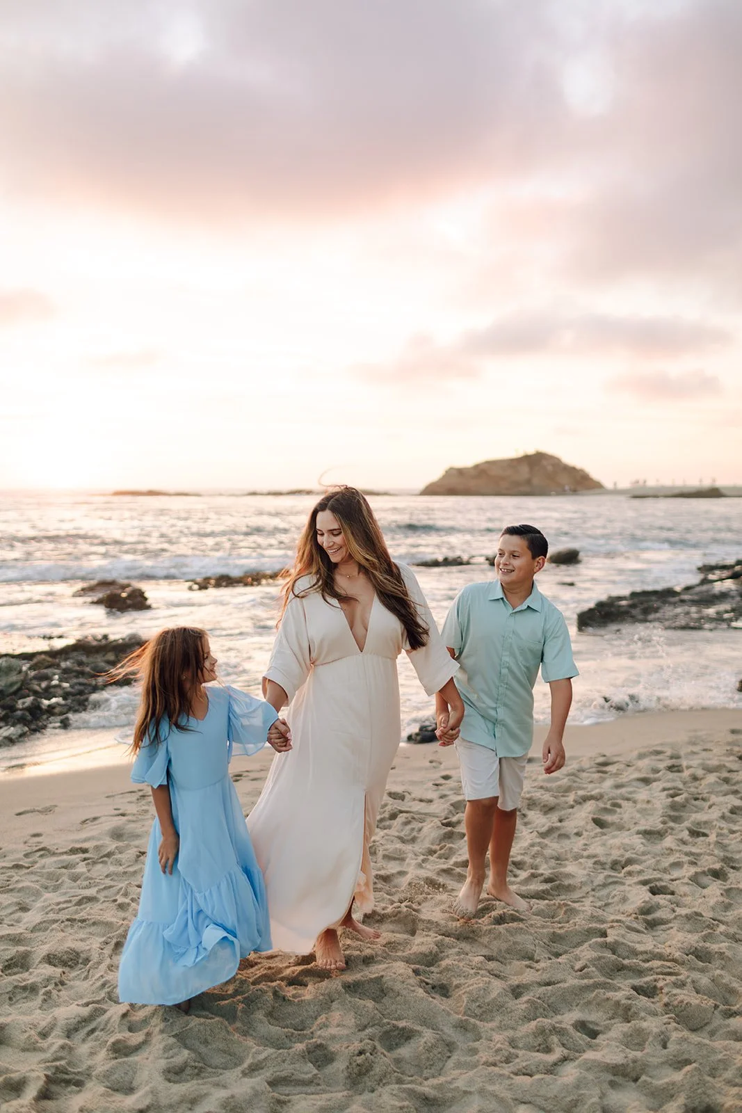 A woman and two children walking hand in hand on a beach at sunset, smiling and enjoying the moment.