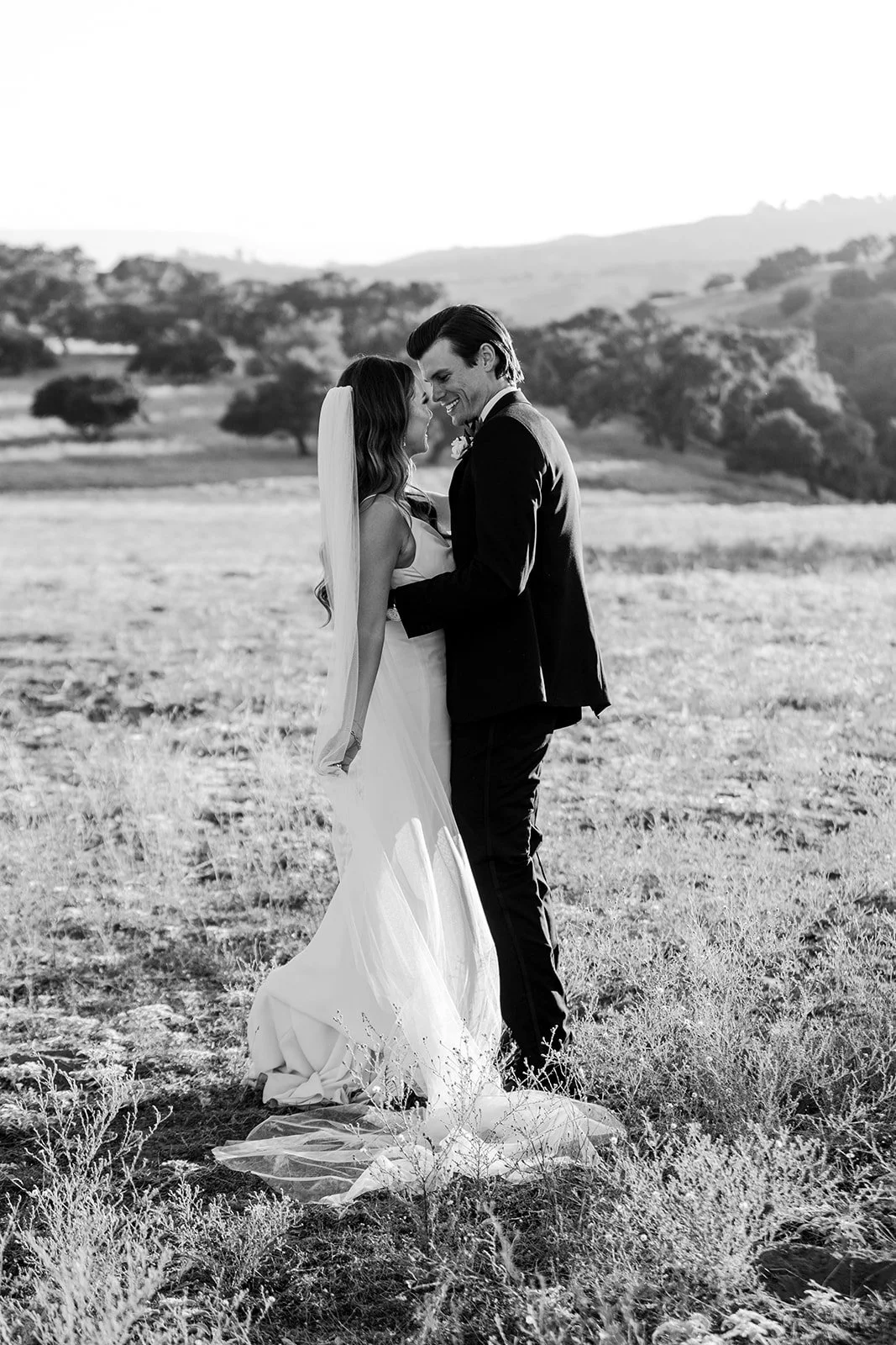 A bride and groom smiling and touching foreheads outdoors in a field with trees and hills in the background, black and white photograph.