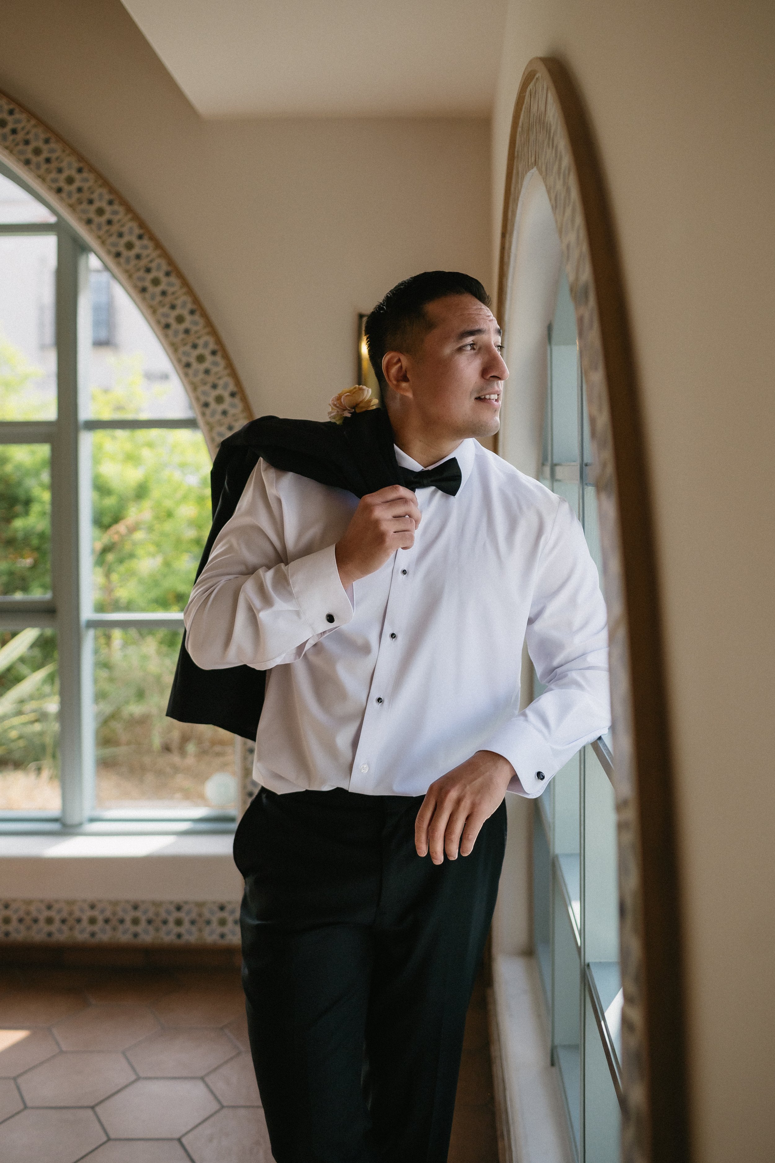 A man in a white tuxedo shirt and black pants looking out a window, holding a black jacket with a pink flower over his shoulder, in a well-lit room with a decorative mirror and hexagonal tile flooring.