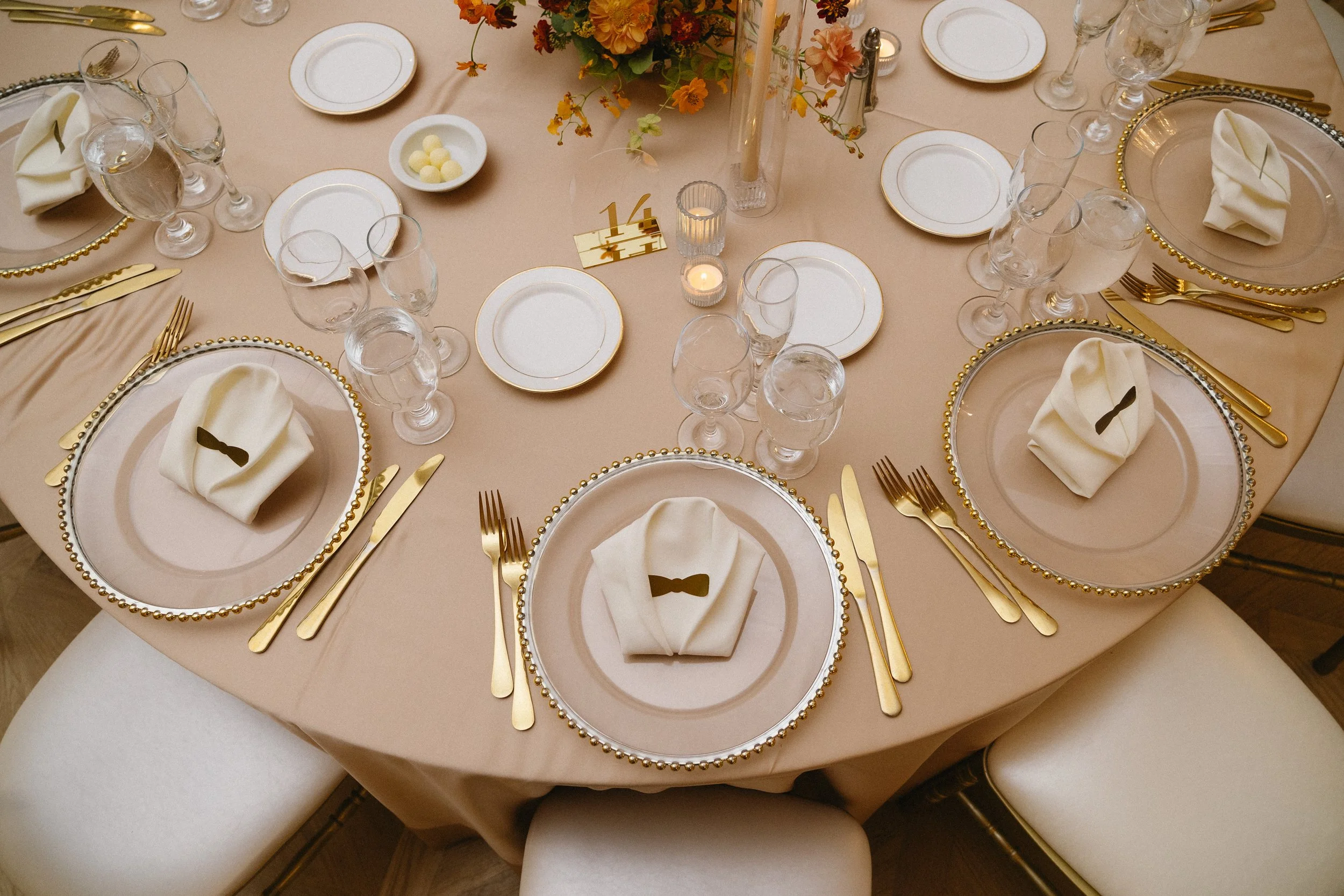 Elegant round dining table set for a formal meal with gold and white place settings, white napkins with black ribbon accents, multiple glasses, a floral centerpiece, and table number 14.