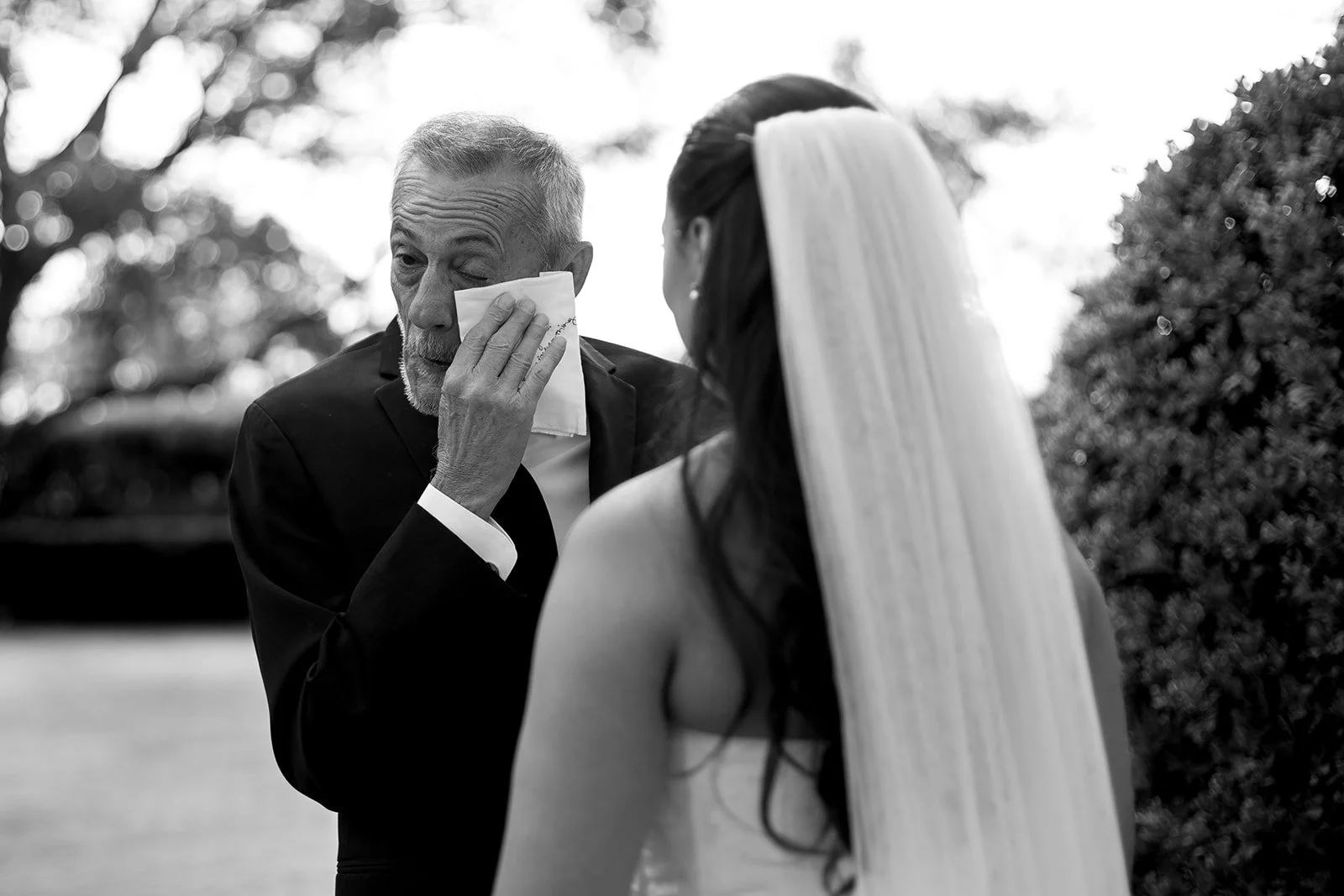 An elderly man with a tissue appears to be crying during a wedding ceremony, with a bride in a white dress and veil facing him outdoors.