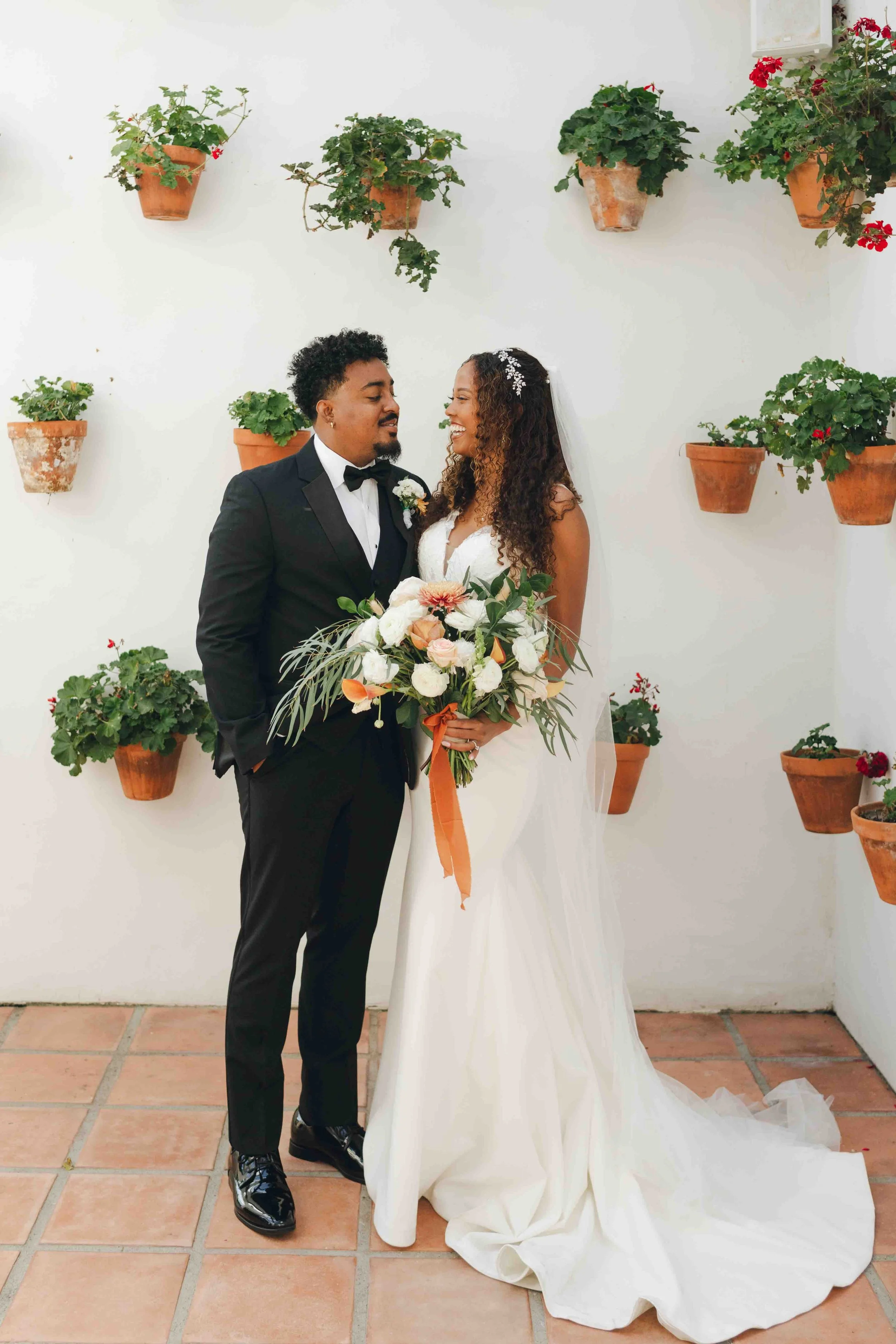 A bride and groom in wedding attire look at each other, with the bride holding a large bouquet of white and pink flowers. They are standing on a terracotta tiled floor against a white wall decorated with hanging terracotta pots filled with green plan