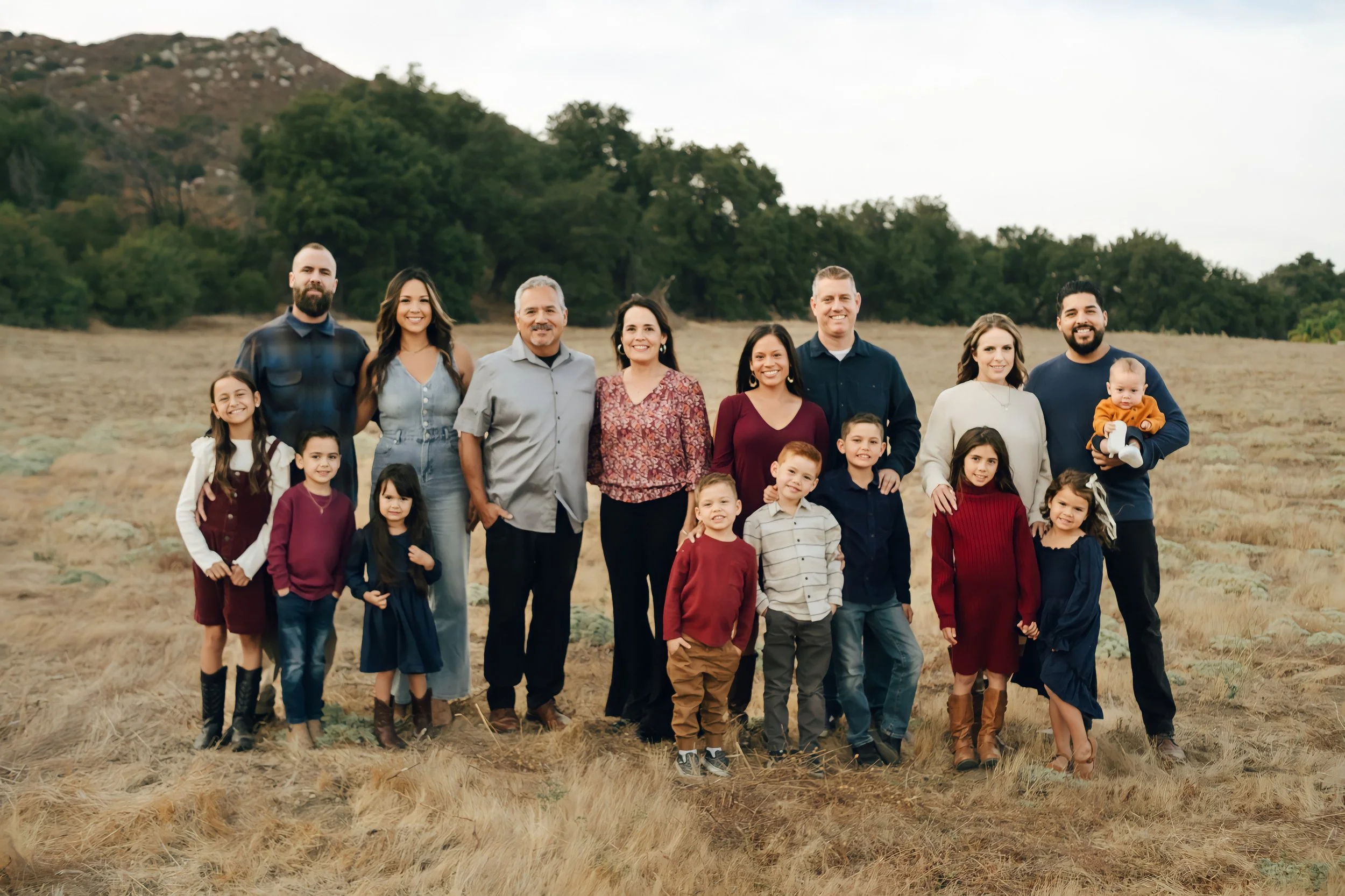 A large multi-generational family standing together outdoors in a field with hills and trees in the background, smiling at the camera.