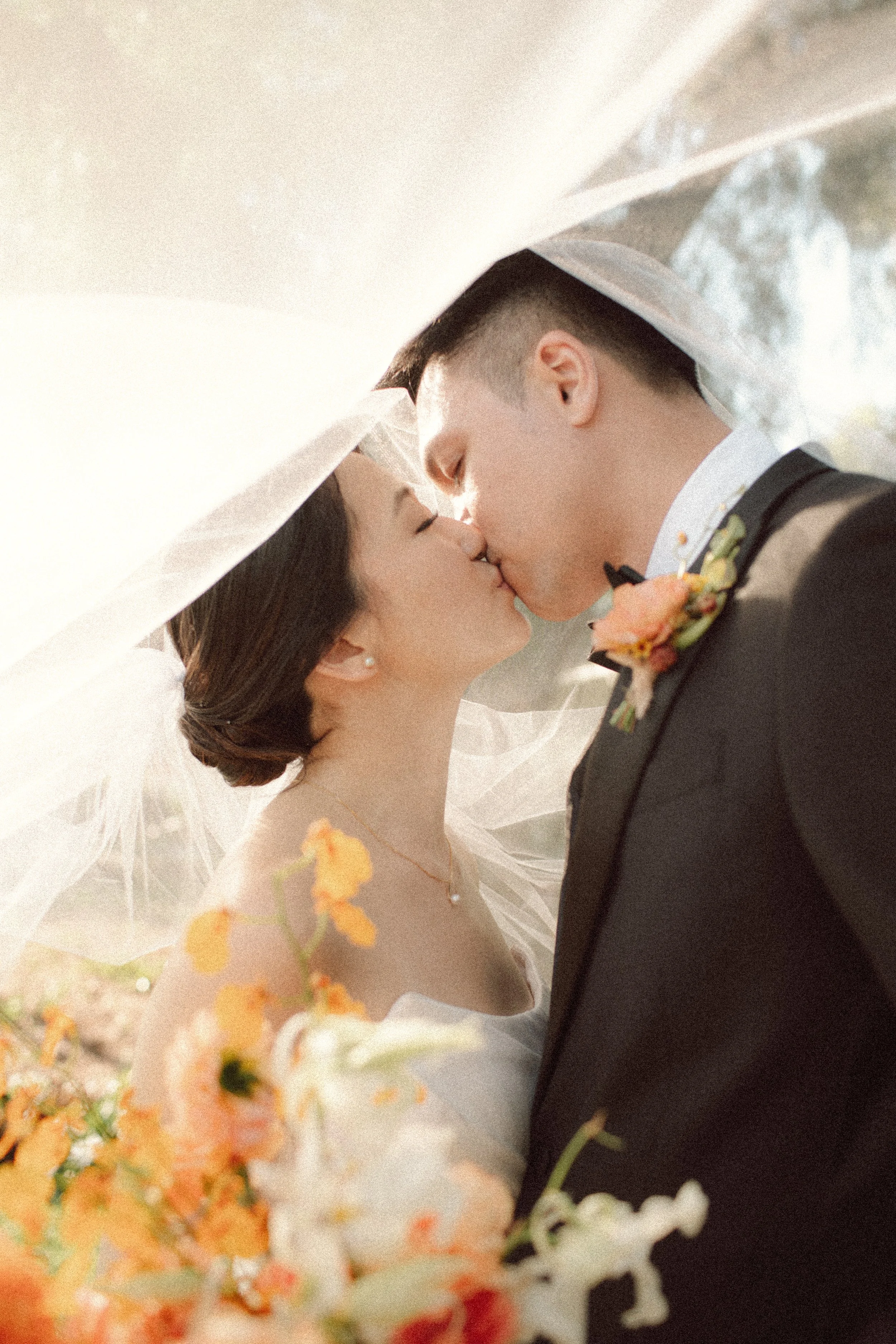 A bride and groom sharing a kiss under a veil outdoors with flowers in the foreground.