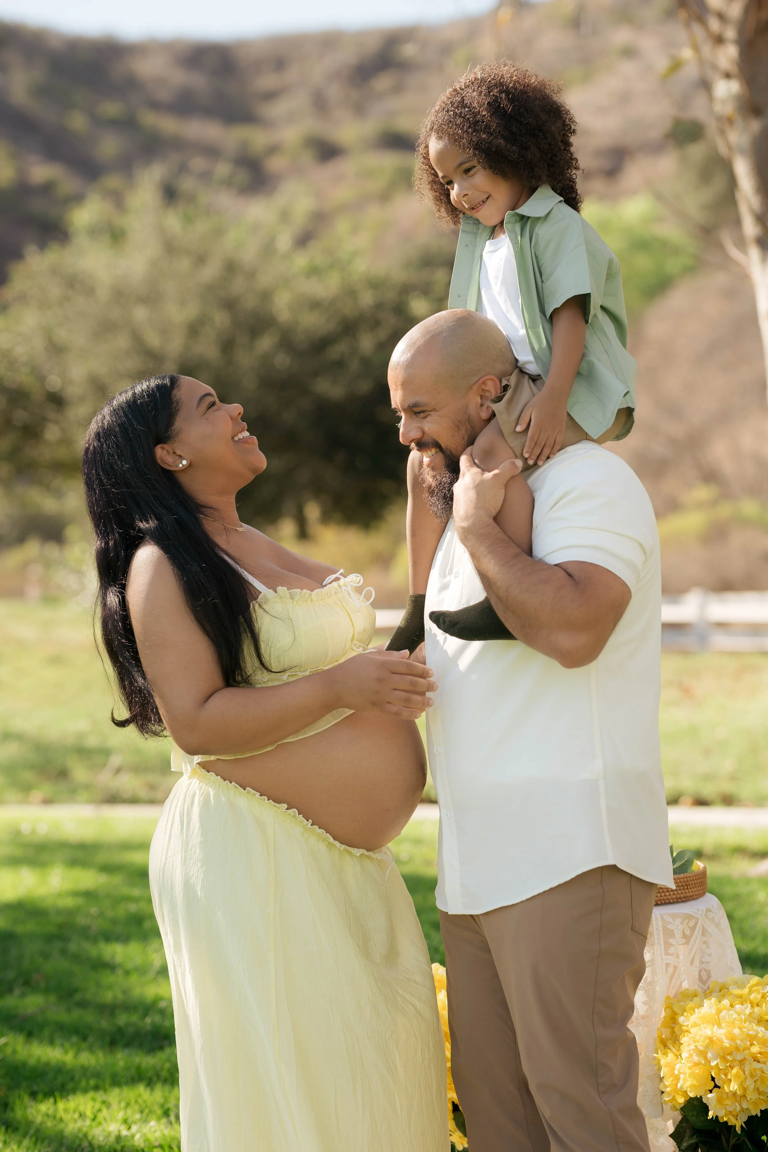 A pregnant woman in a yellow dress and a smiling man holding a young girl on his shoulders outdoors in a park, with green grass, trees, and hills in the background.
