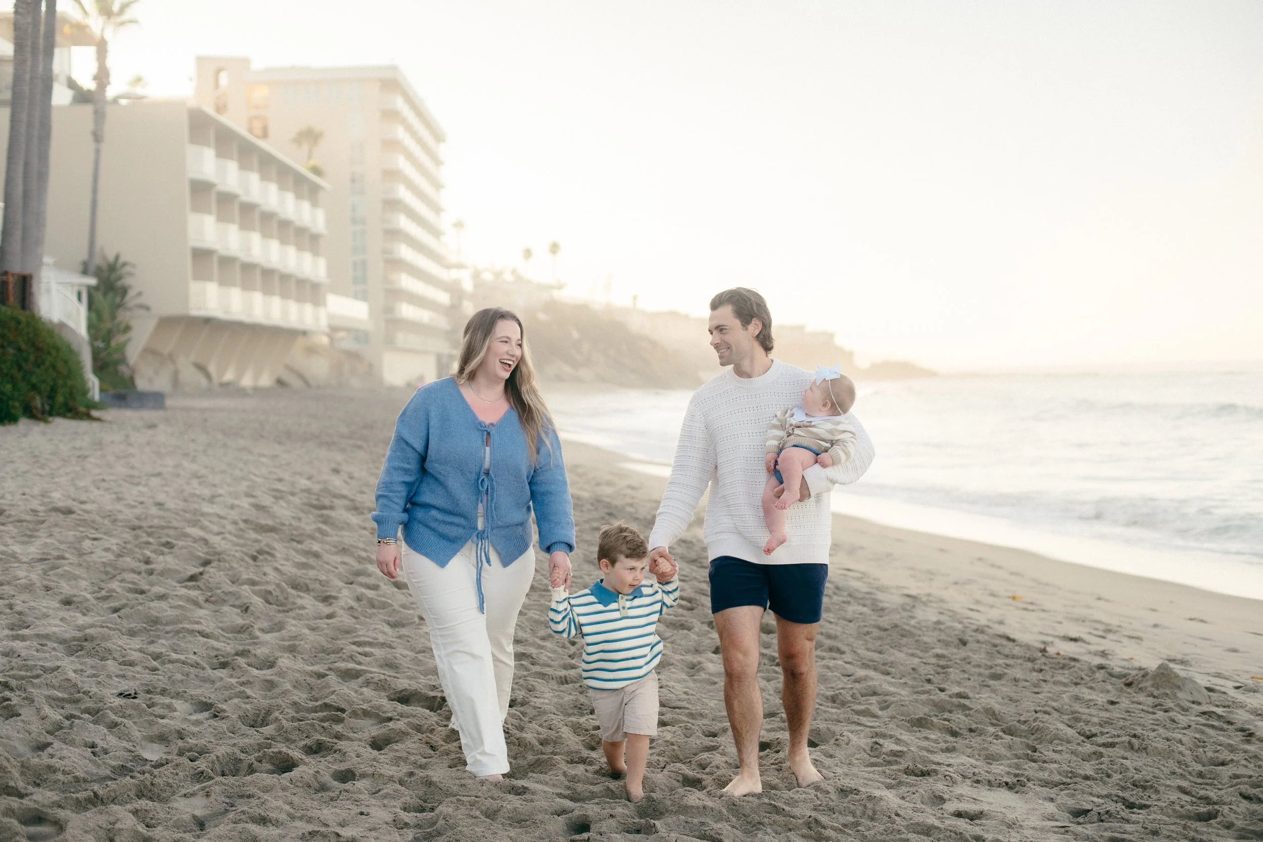 A family walking on the beach during sunset, with two adults, a young boy, and a baby, all smiling and enjoying their time.