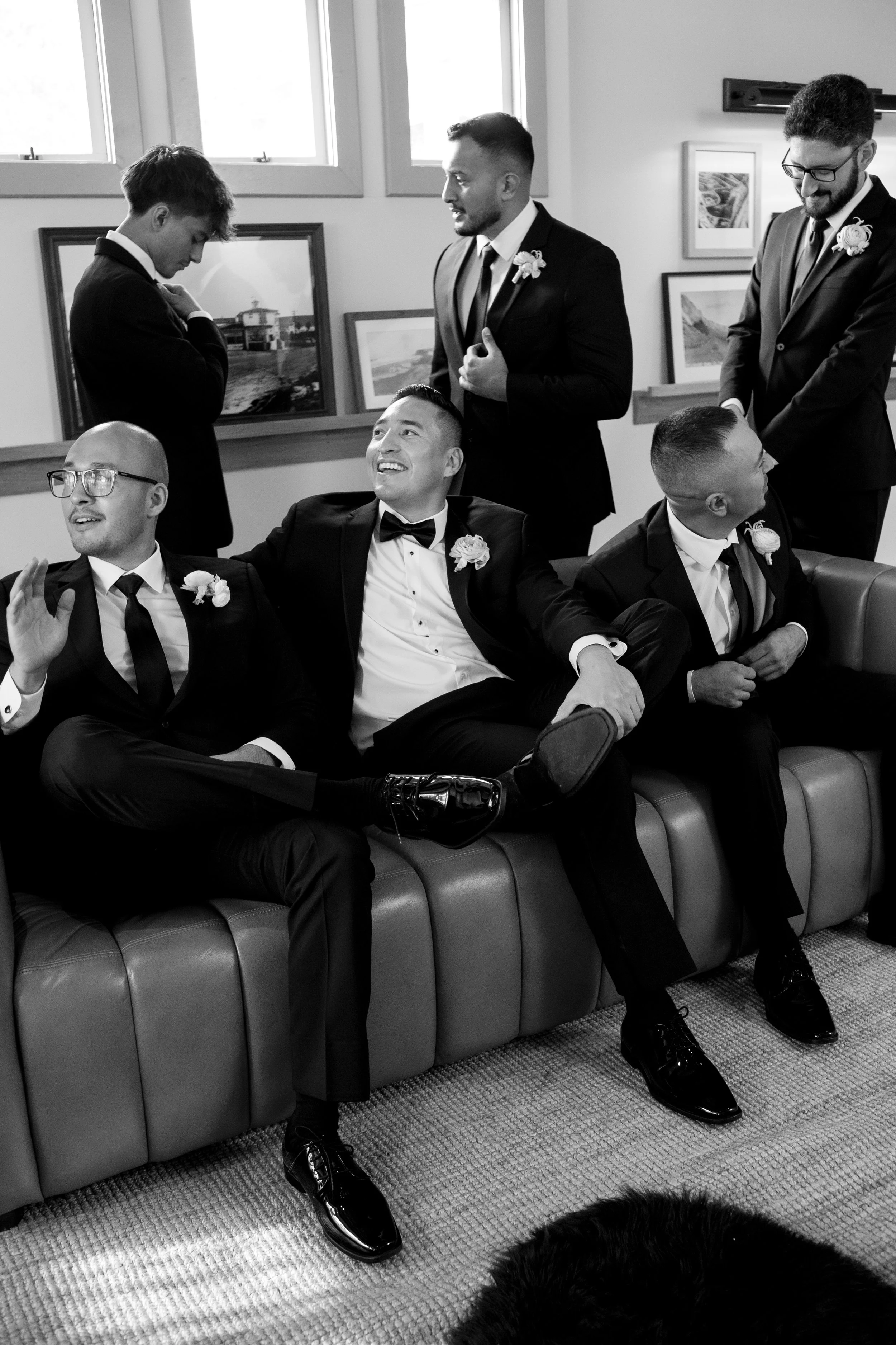 Black and white photo of six men dressed in tuxedos, sitting and standing inside a room with framed pictures on the wall. They are smiling and engaged in conversation, likely at a wedding or formal event.