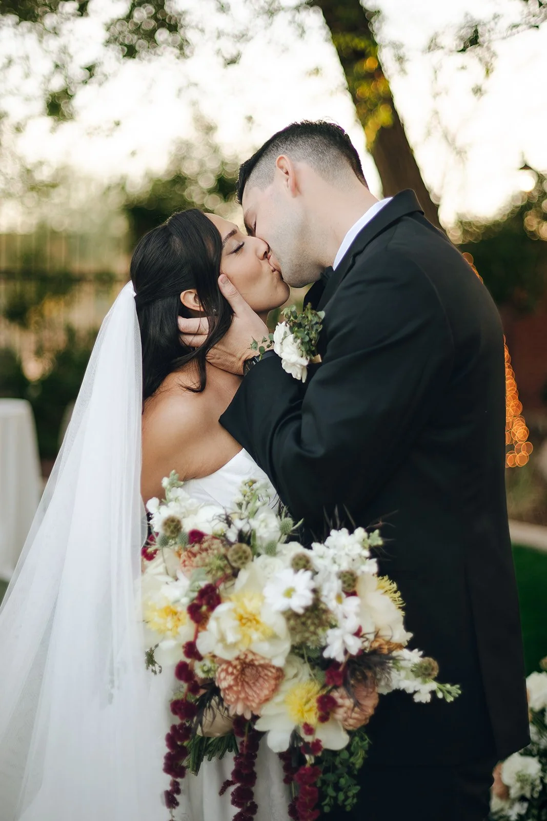 A bride and groom kissing outdoors, with the bride holding a large bouquet of flowers, during sunset.
