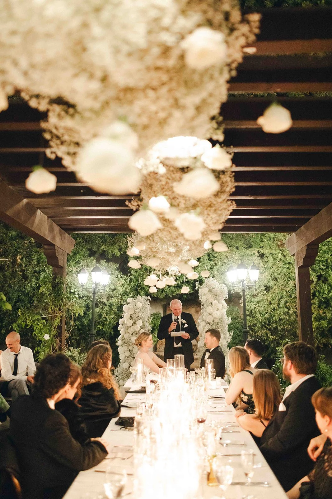 A wedding reception is taking place outdoors under a pergola decorated with hanging white flowers and floral arrangements. Guests are seated at a long table set with glasses and candles. A man in a tuxedo is giving a speech, while the bride and groom