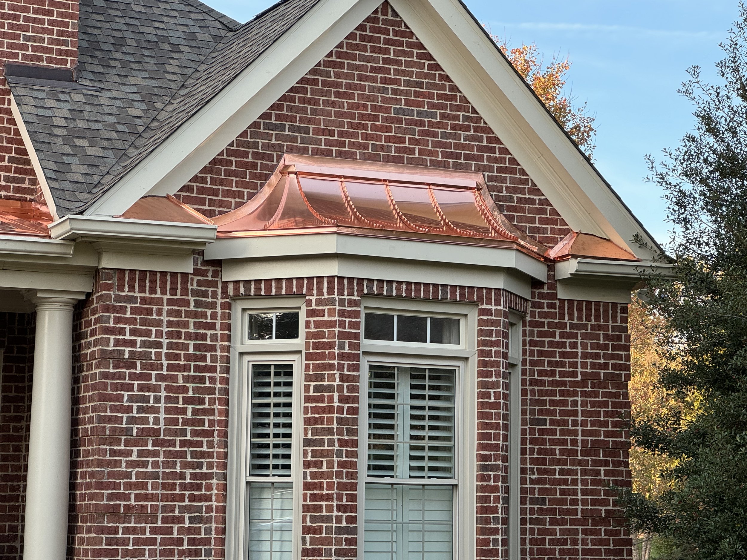 Close-up of a brick house corner with a copper roof section and white window shutters, surrounded by trees with fall foliage.