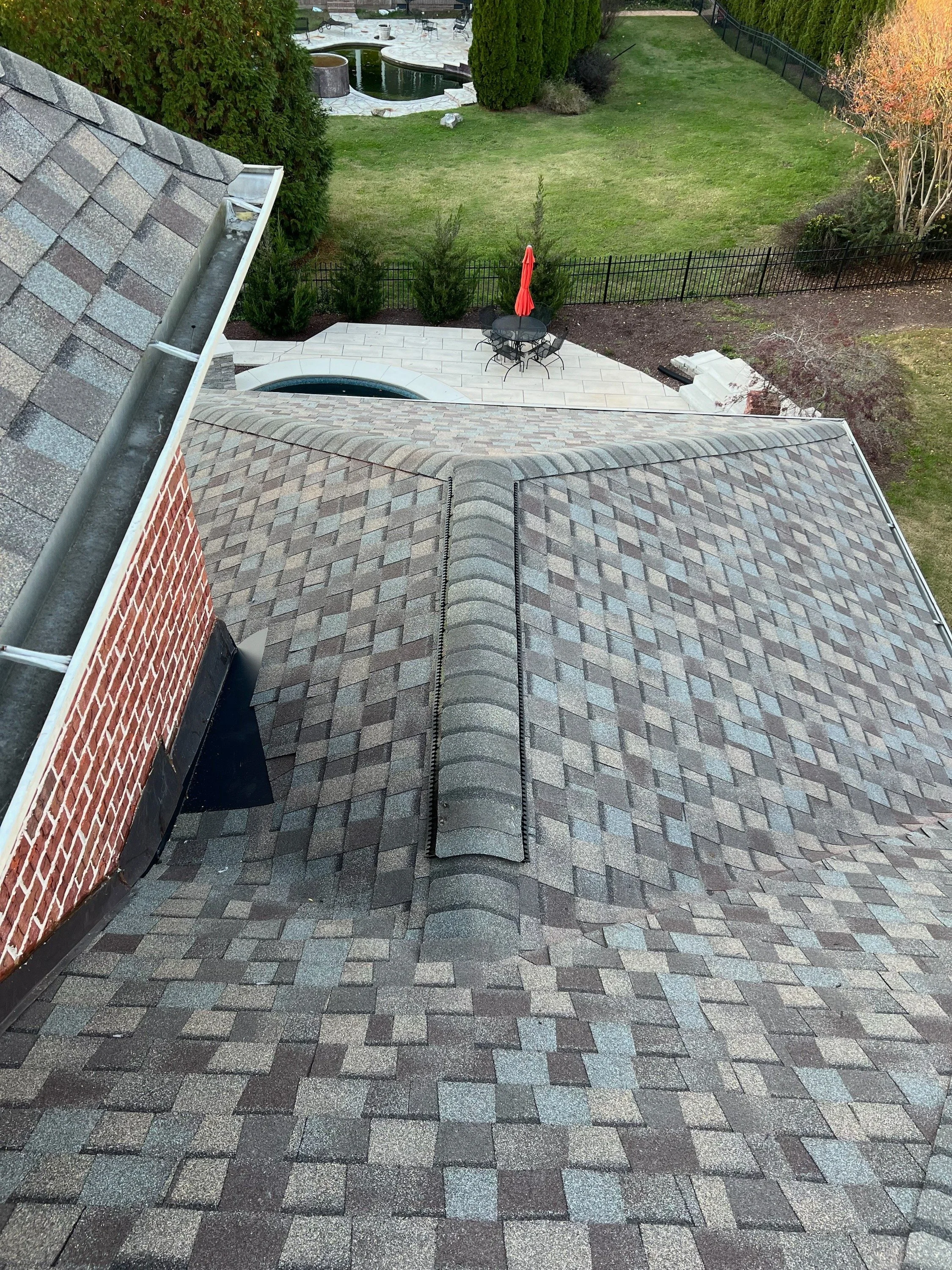 View from a roof showing asphalt shingles, a brick chimney, and a vent pipe. Below, there is a backyard with a patio table, chair, umbrella, a small pool, trees, and a grassy lawn.
