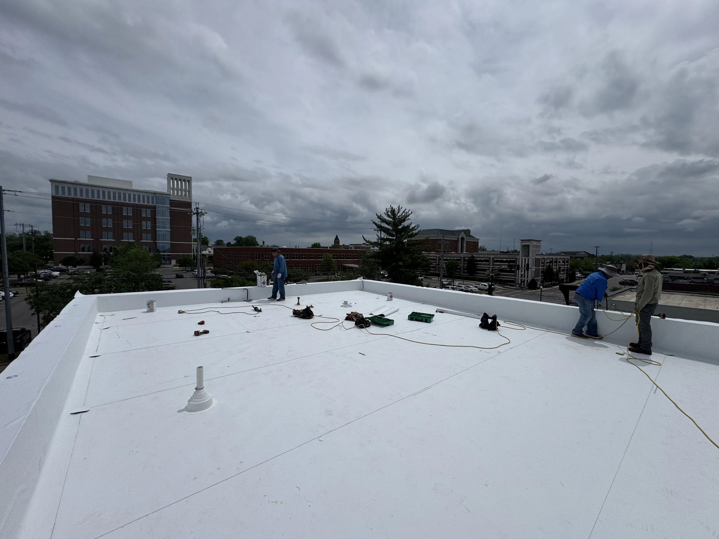 Workers on a rooftop installing or repairing a white roofing surface under a cloudy sky.