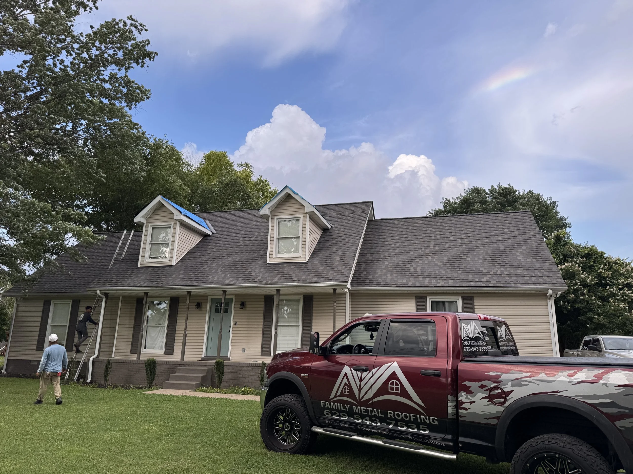 Two workers are installing a metal roof on a house, with one climbing a ladder and another standing on the roof. A pickup truck with the logo 'Family Metal Roofing' is parked in front of the house.
