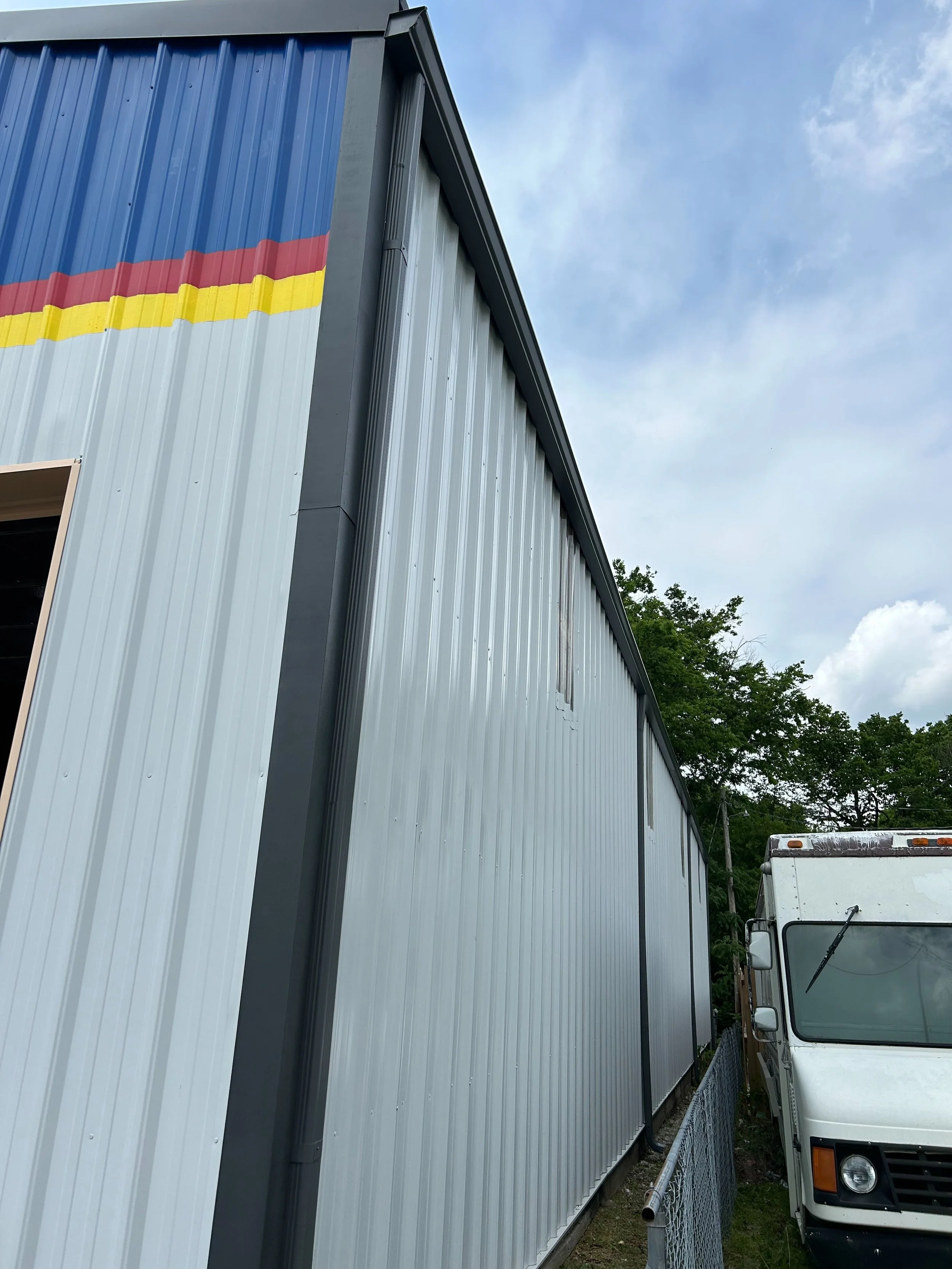 Side view of a large metal building with a colorful stripe near the top, a white truck parked beside it, and trees in the background under a cloudy sky.