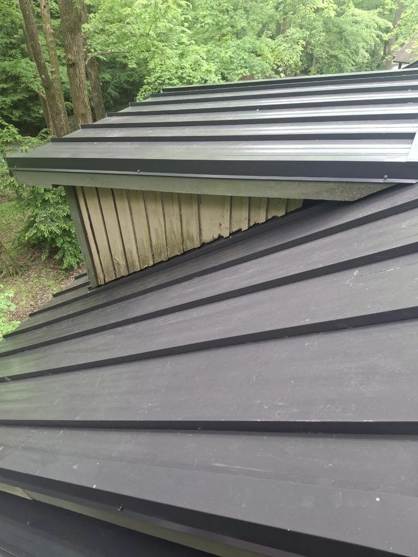 A close-up view of a metal roof on a building, with a wooden dormer protruding from the roof. Green trees are visible in the background.