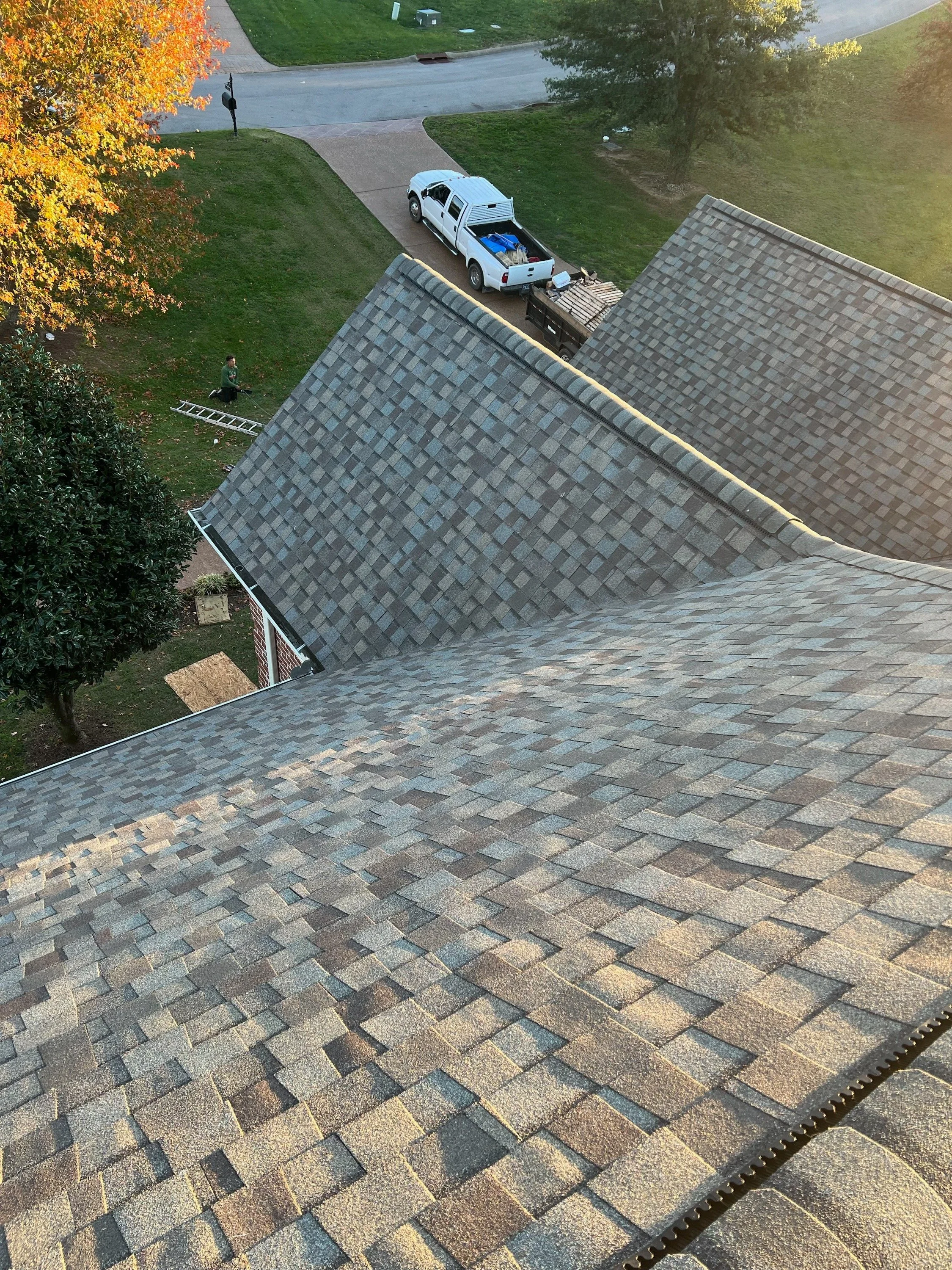 View from the roof of a house showing asphalt shingles, a yard with a tree and a person working, and a street with a parked pickup truck and some equipment in the driveway.