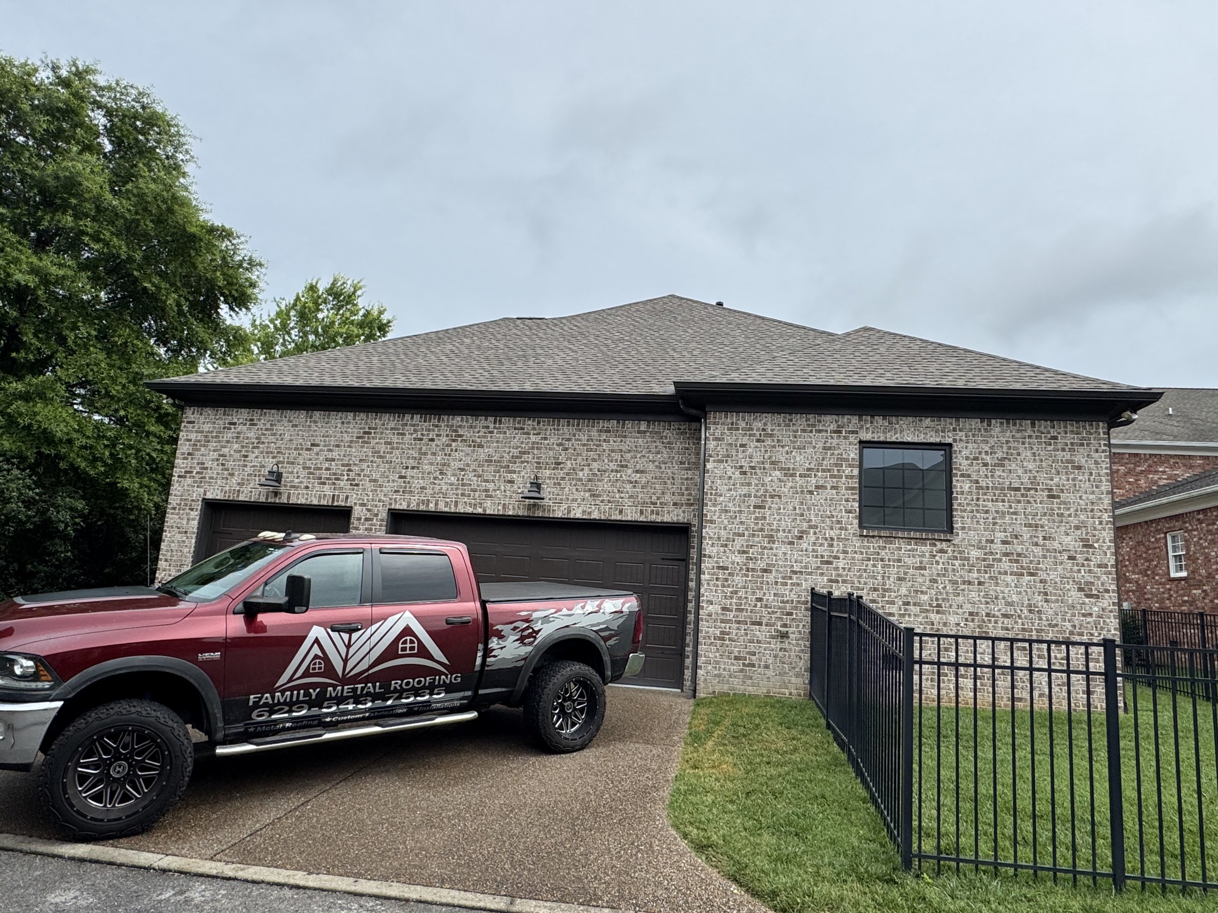 A modern brick house with a sloped roof, black garage door, a window, and a driveway. A red truck with black wheels, advertising "Family Metal Roofing" and a phone number, is parked in front.