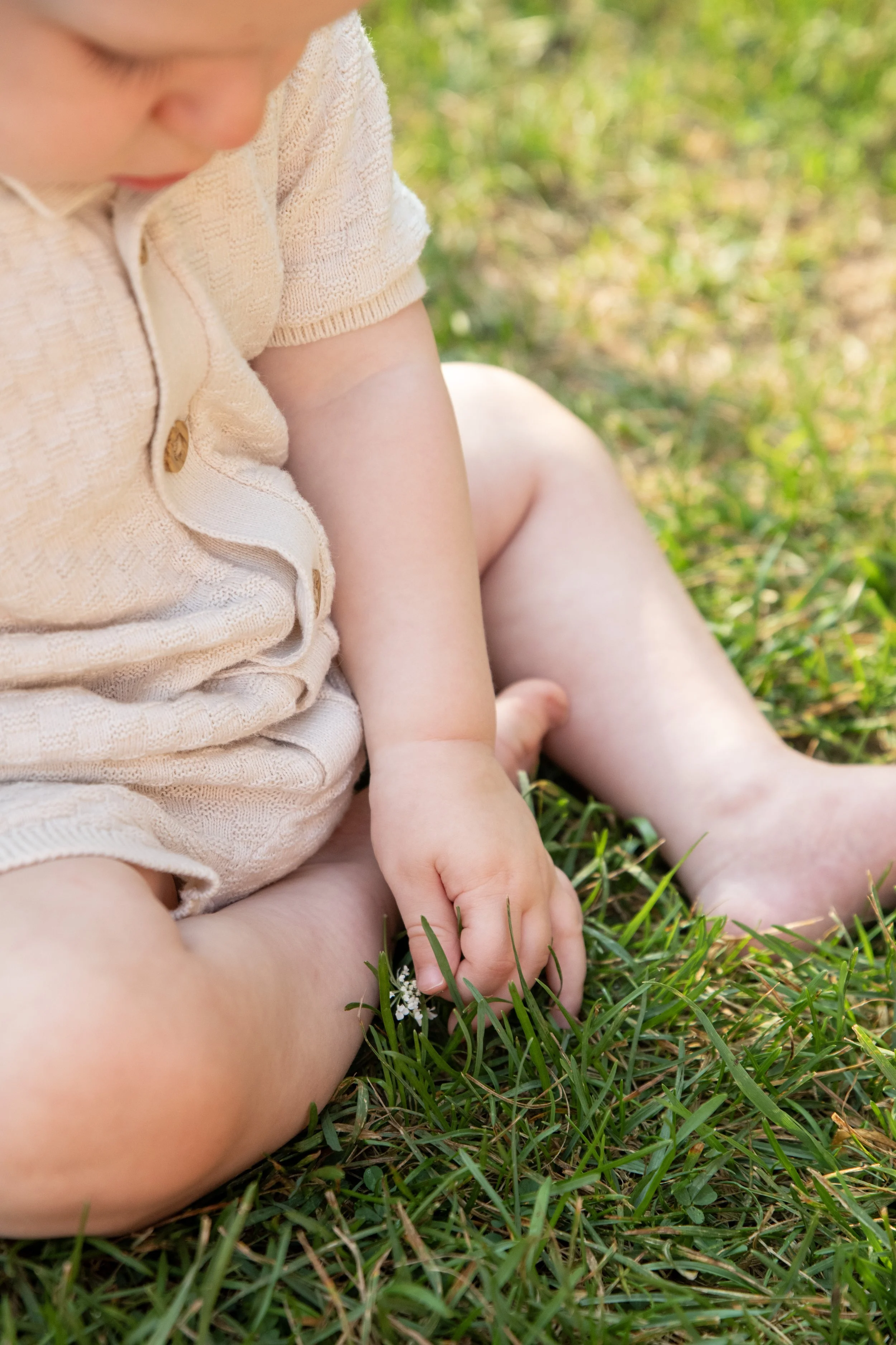 A close-up of a young child sitting on grass outside, wearing a beige knitted outfit. The child is touching the grass with one finger, with blurred greenery in the background.