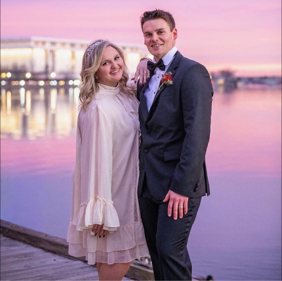 A happy couple dressed in wedding attire, standing on a dock near water during sunset, with a bridge in the background.