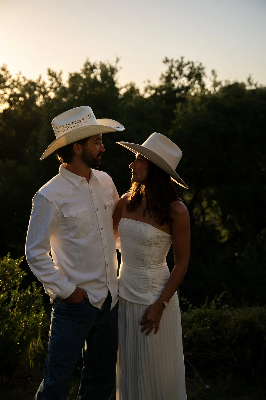 A man and a woman wearing cowboy hats and casual clothing standing close together outdoors at sunset, looking into each other's eyes.