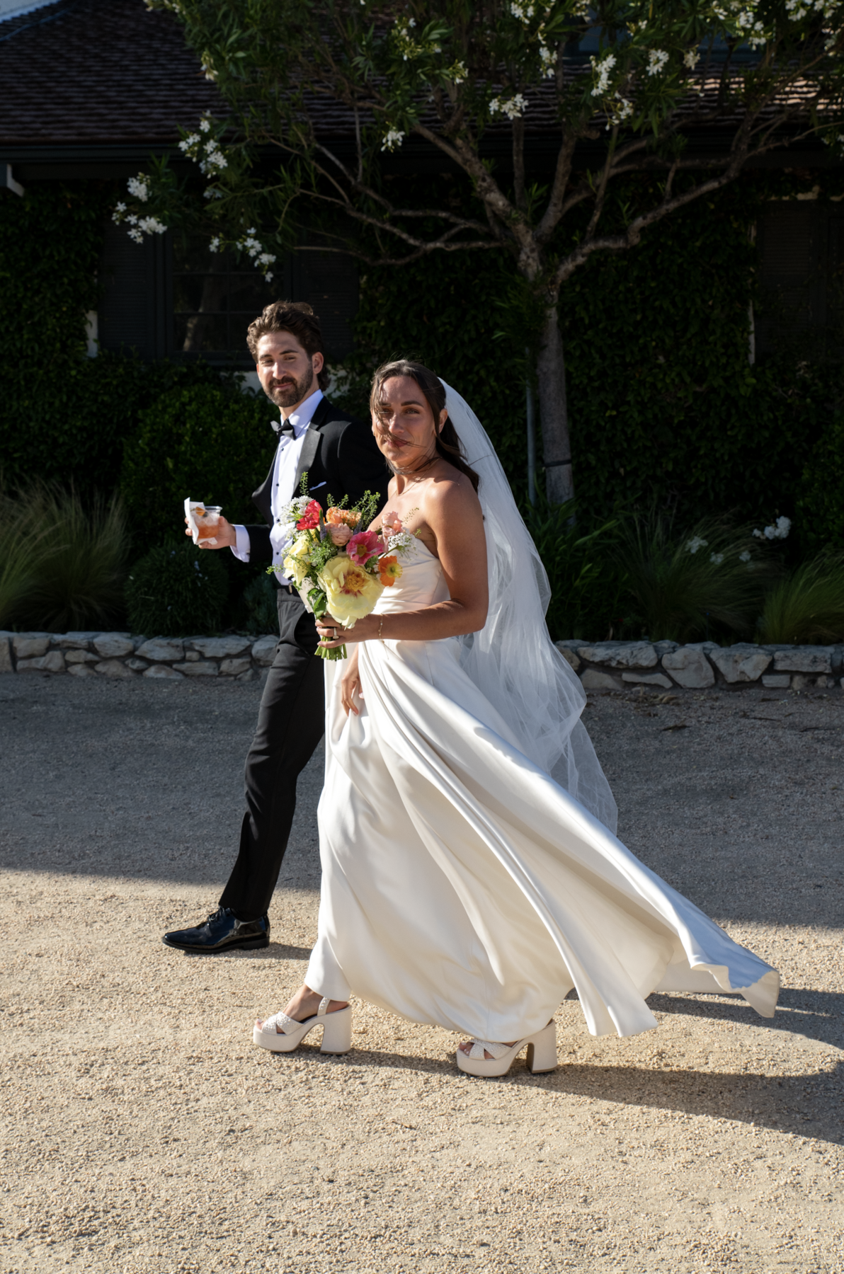 A bride and groom walking outdoors on a sunny day. The bride is holding a bouquet and wearing a strapless white wedding dress with a veil. The groom is dressed in a black tuxedo with a bow tie. They are smiling and walking along a gravel path, with g