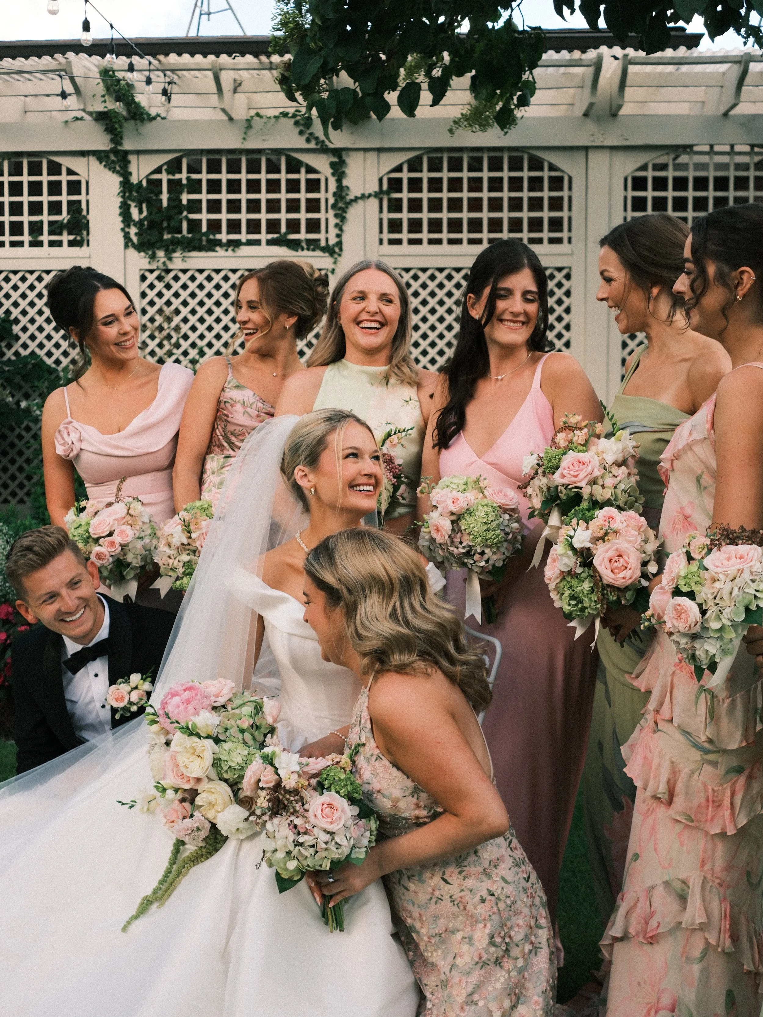 A bridal party at a wedding reception, including women in floral dresses and a bride holding a bouquet, smiling and laughing together, with a white lattice fence and string lights in the background.