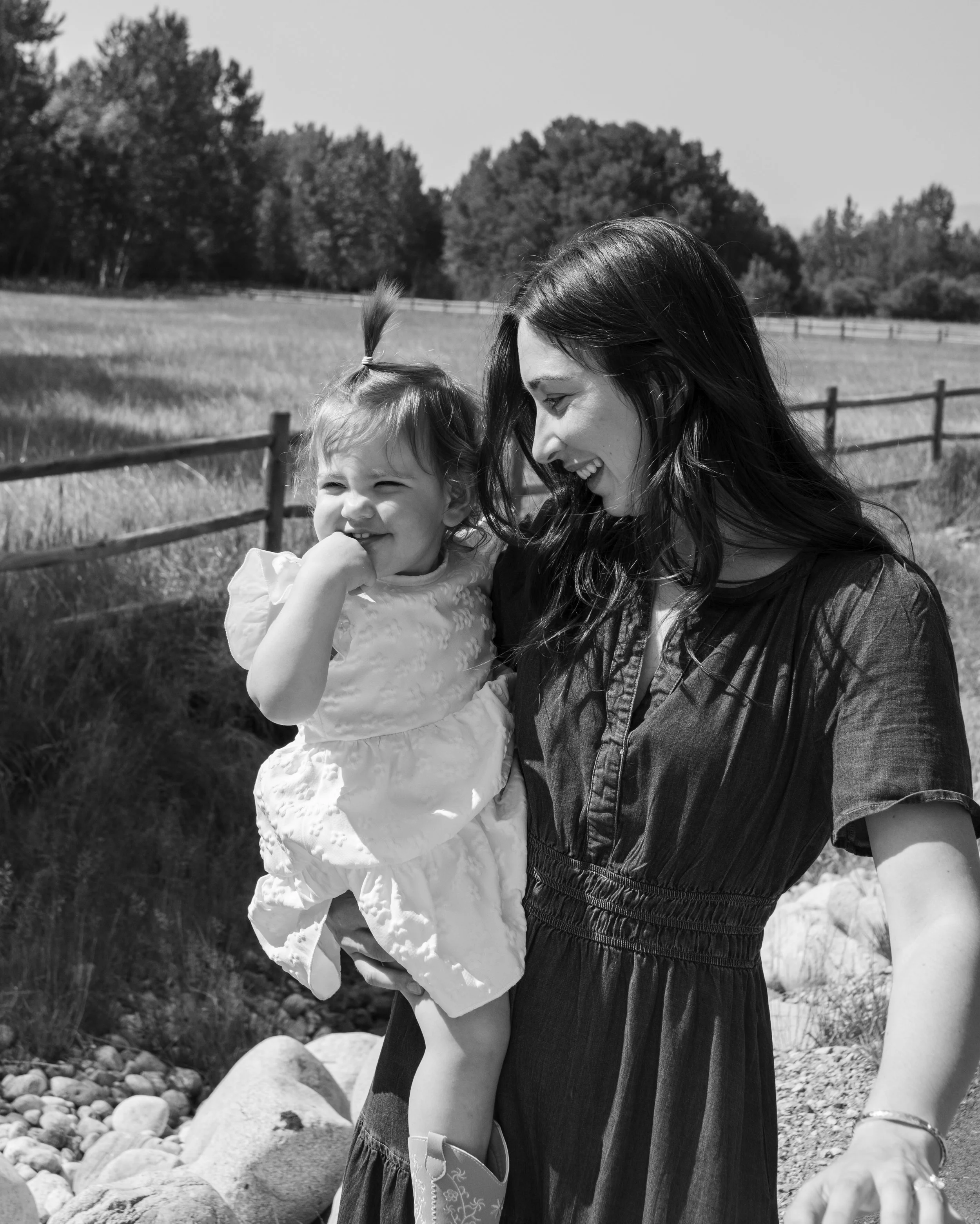 A woman with long dark hair smiling and looking at a little girl with a ponytail, outdoors in a grassy area with rocks and trees in the background.