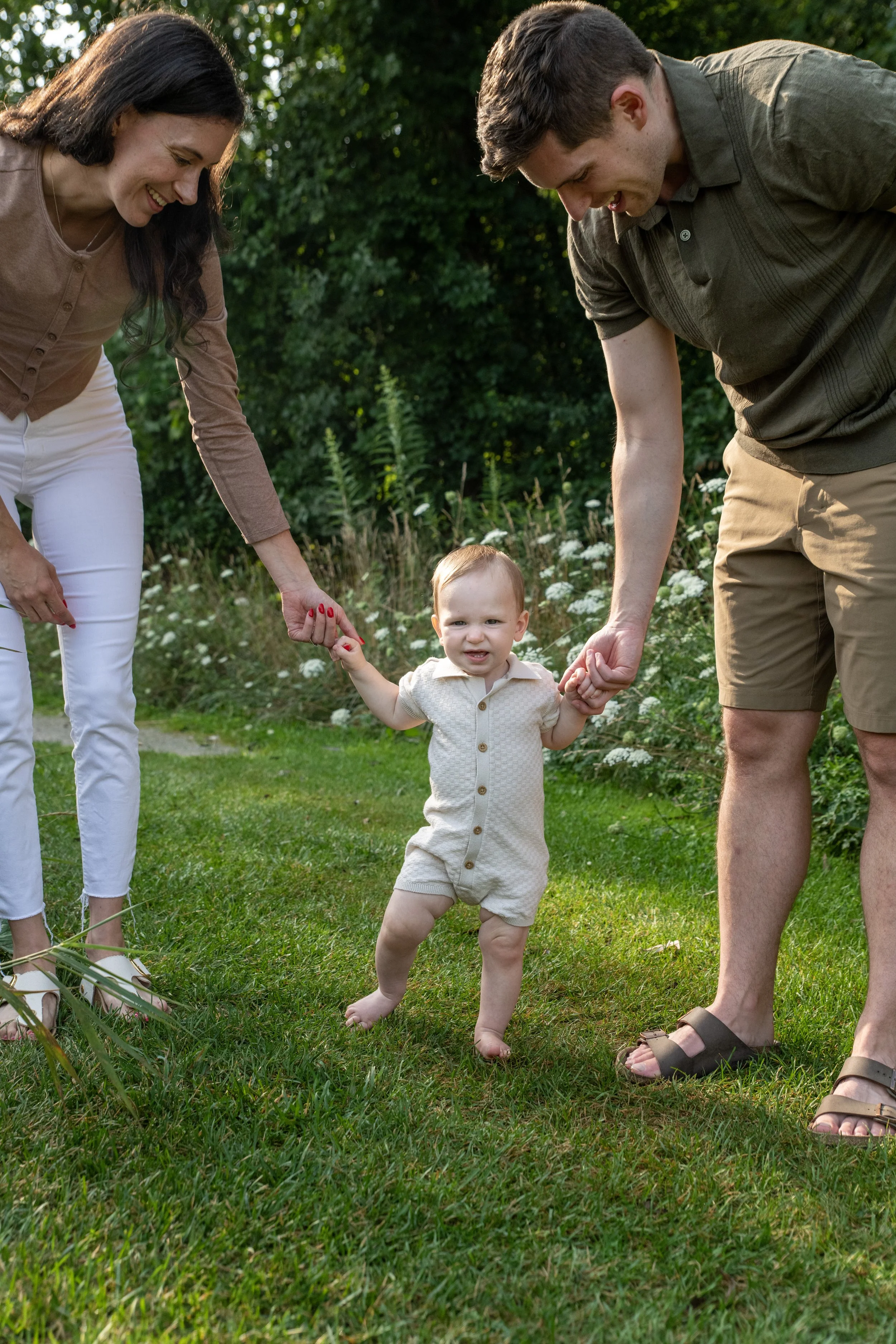 A family of three, including a mother, father, and their toddler, holding hands and walking through a grassy outdoor area with trees and white flowers in the background. The mother is smiling and dressed in white pants and a brown top, the father is 