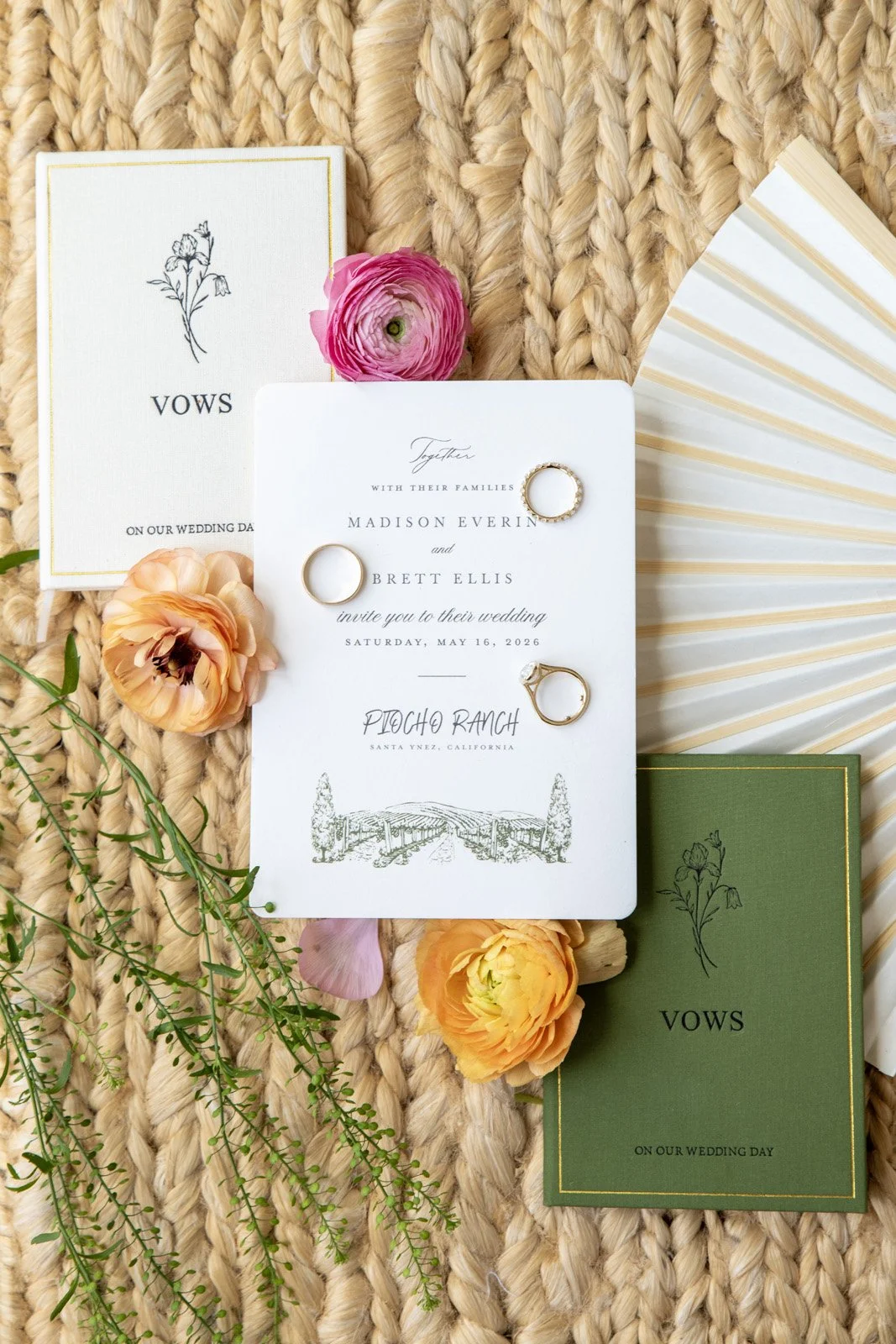 Wedding invitation suite with rings, flowers, and greenery on a woven background.