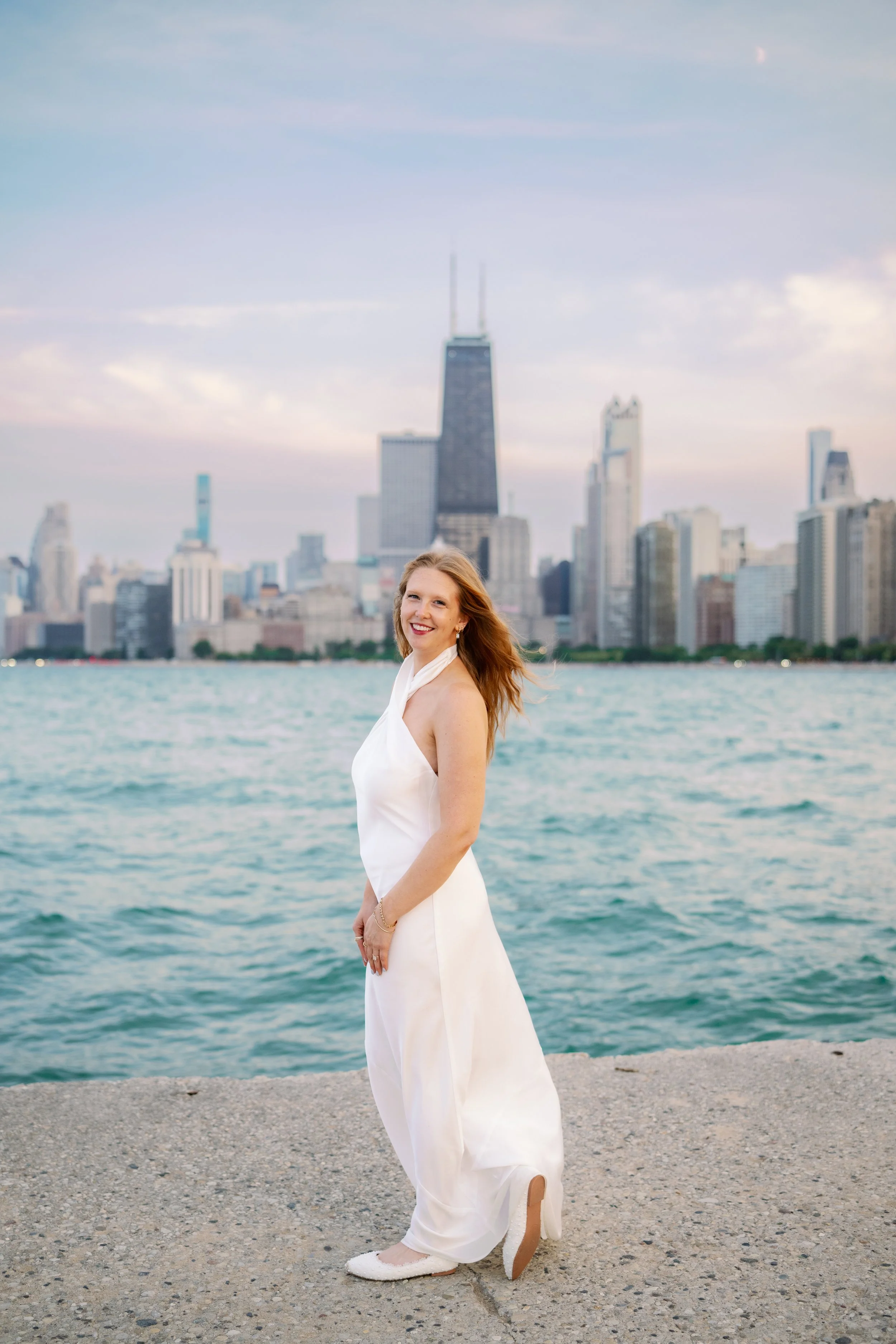 A woman in a white dress standing on a shoreline with water and a city skyline including a tall building with antennas in the background.