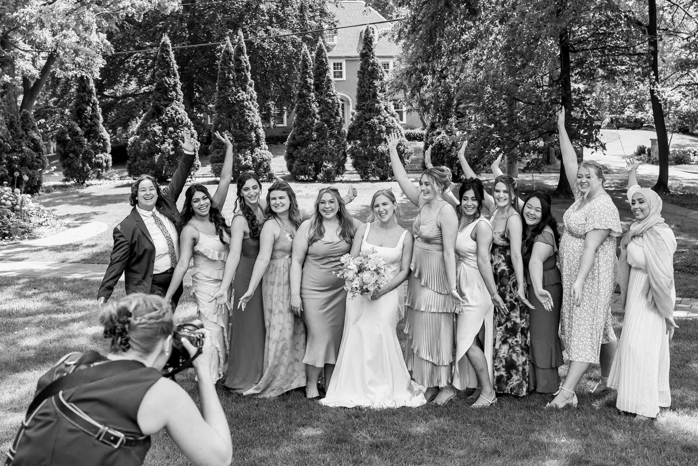 A group of women in formal dresses and a suit, celebrating outdoors in a garden with trees and a house in the background. A photographer is taking a picture of them.