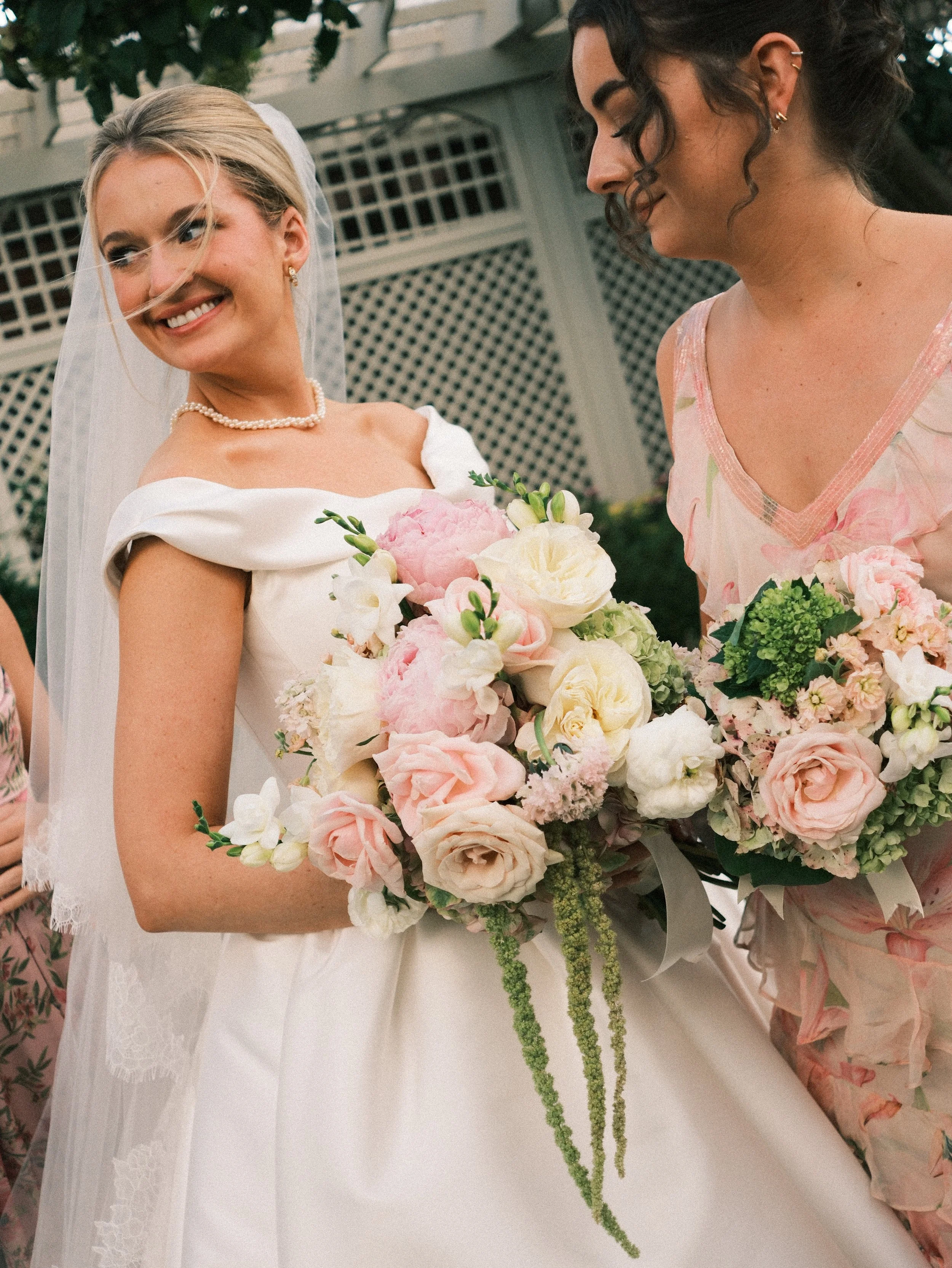 Two women at a wedding, holding bouquets of flowers and smiling at each other. One woman is dressed as a bride in a white gown with a pearl necklace and veil, the other is in a pastel pink dress with floral patterns.